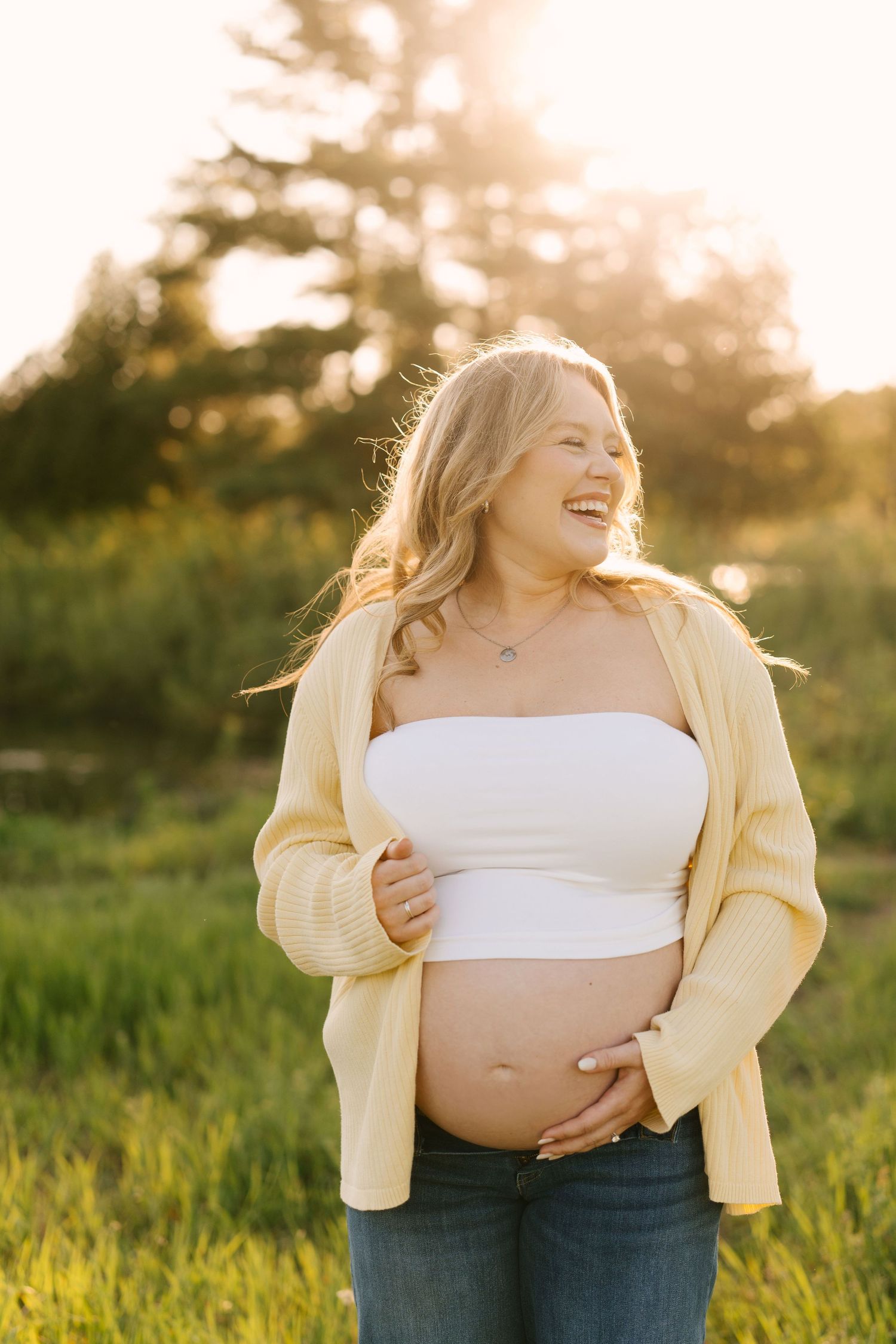 Laughing pregnant woman in yellow cardigan and white tube top holds her baby bump outdoors at golden hour.