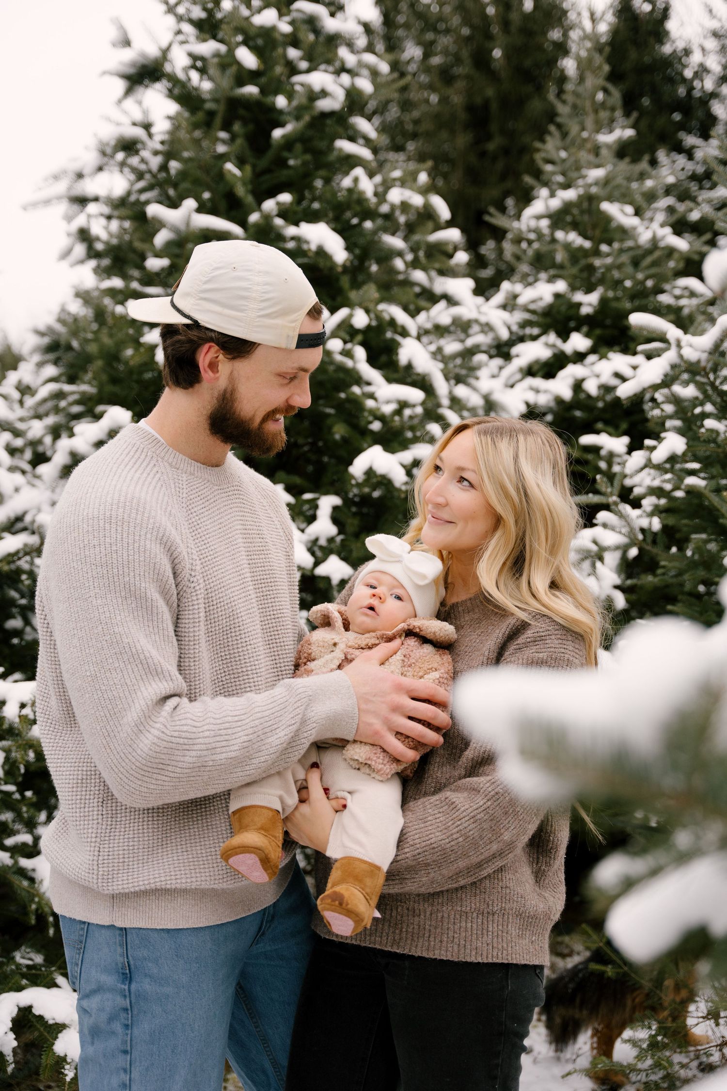 Happy couple holding their baby girl between snow-covered Christmas trees at a winter tree farm.