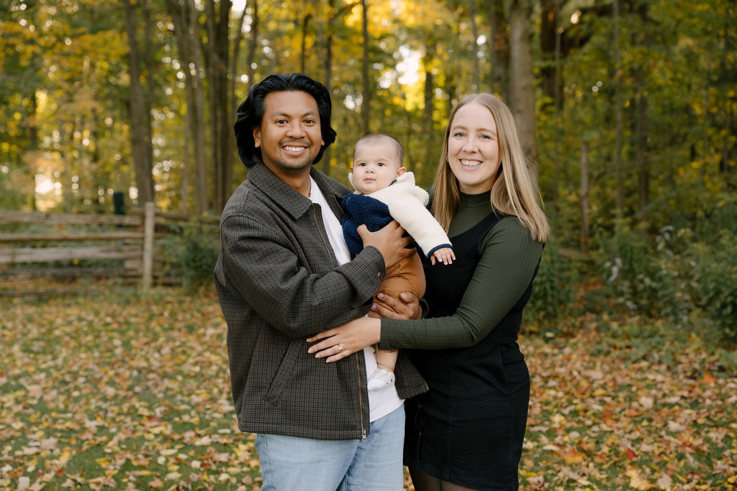 Interracial family smiling in autumn park, parents holding baby among colorful fall foliage.