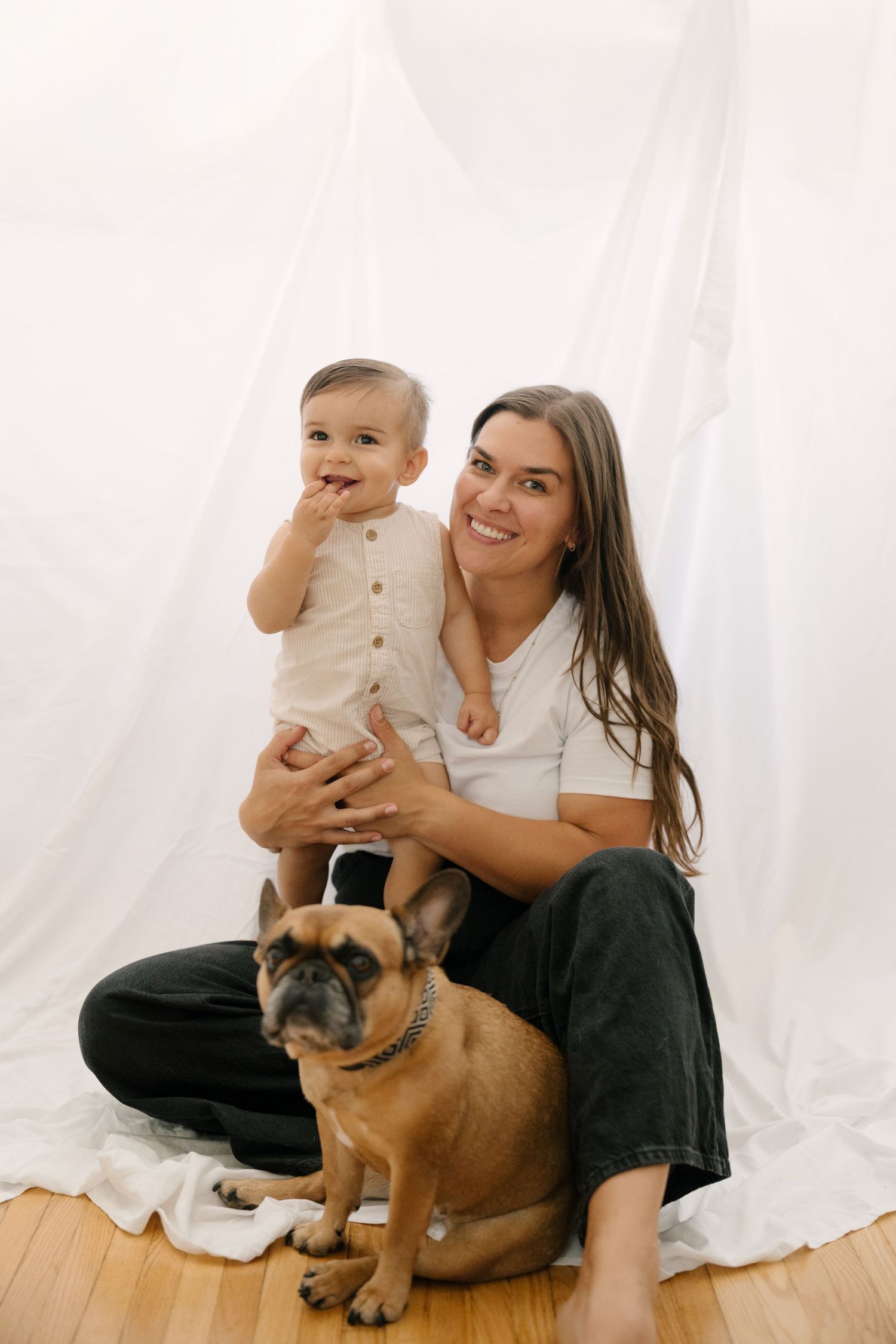 Smiling mother holds her baby boy beside their French Bulldog while posing against a white backdrop.