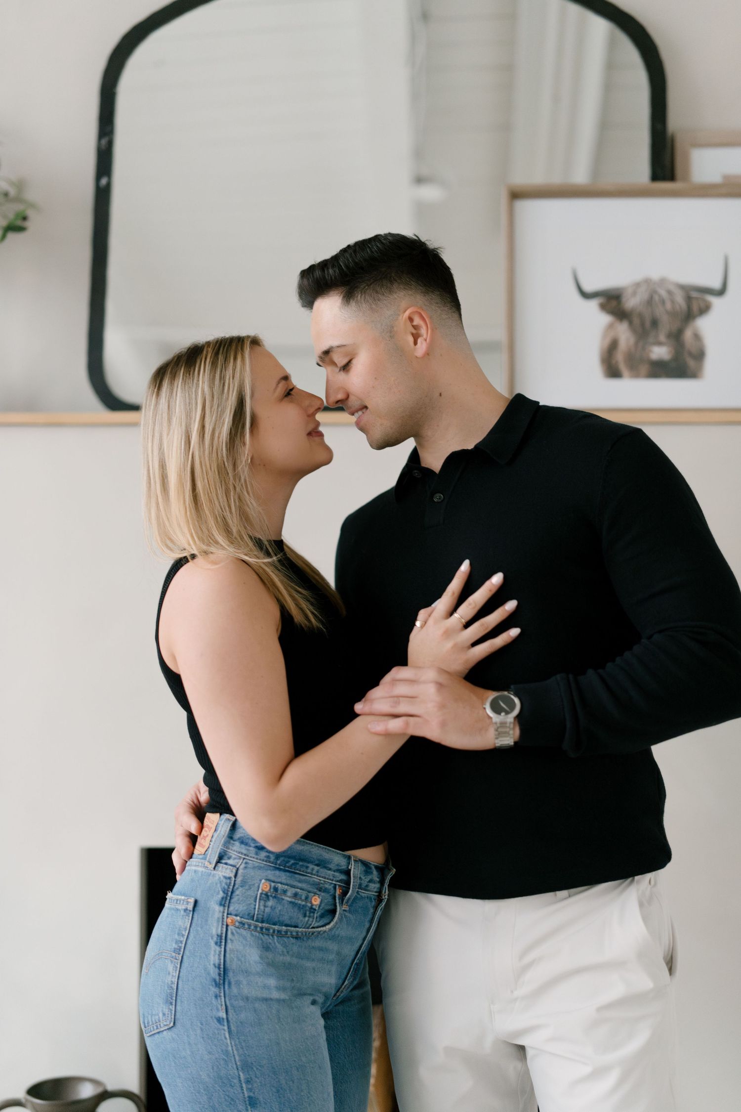 Couple in black outfits leaning in for a kiss in a cozy kitchen with framed wall art.