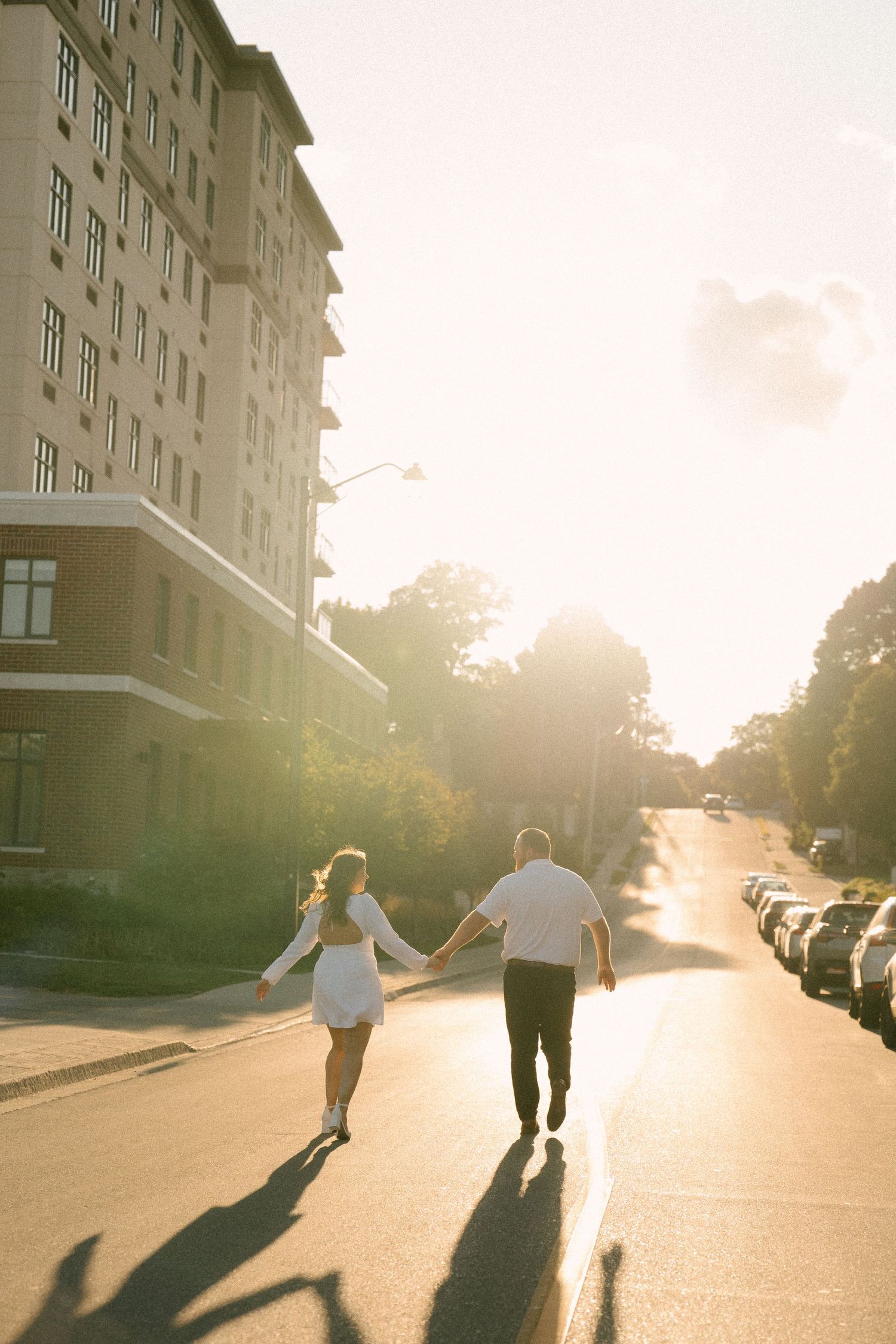 Couple holding hands walking down a sunlit street toward golden sunset light.
