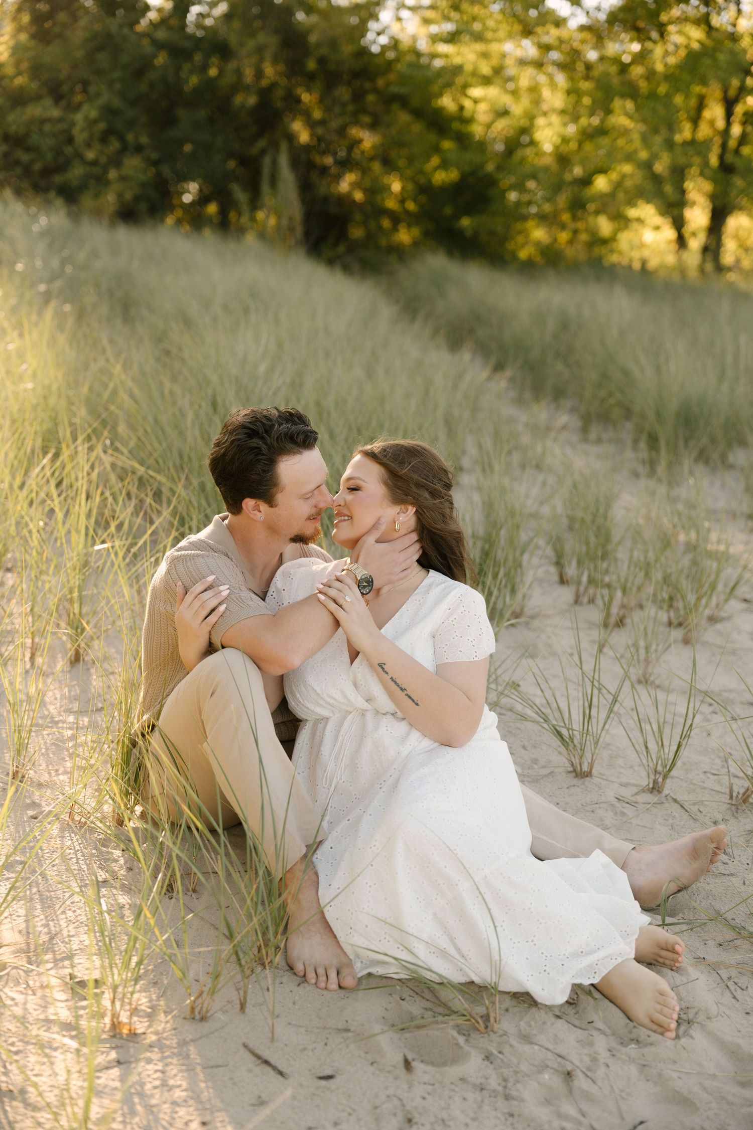 Romantic couple sitting on sand dunes surrounded by tall grass about to kiss.