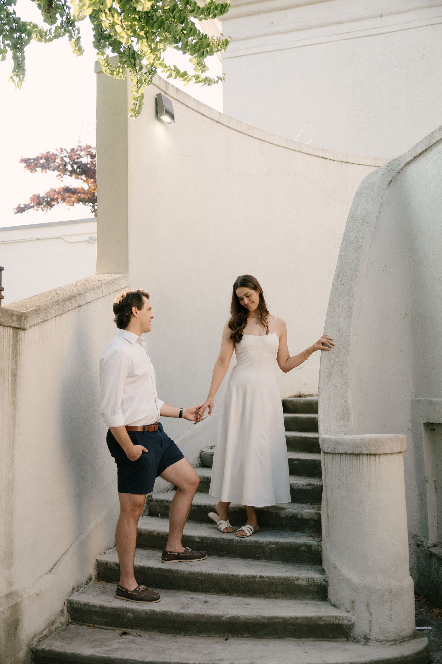 Couple holding hands on elegant curved stone stairs outside a white building.