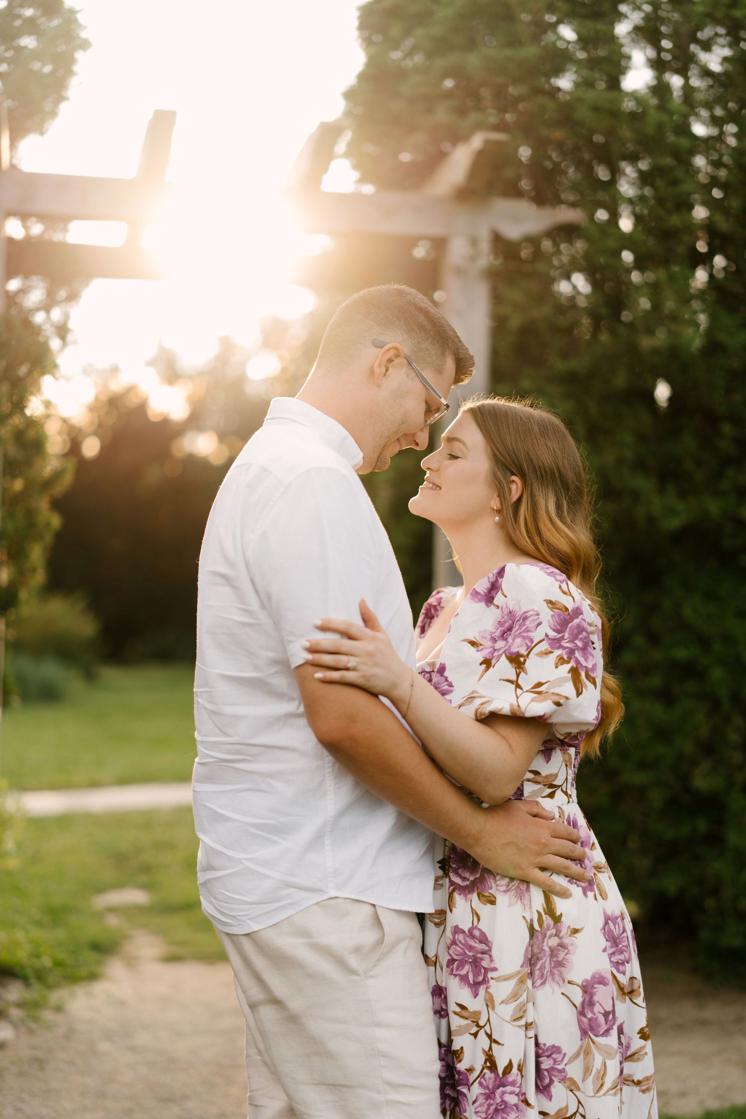 Couple embracing and almost kissing in warm golden sunset light near wooden garden structure.