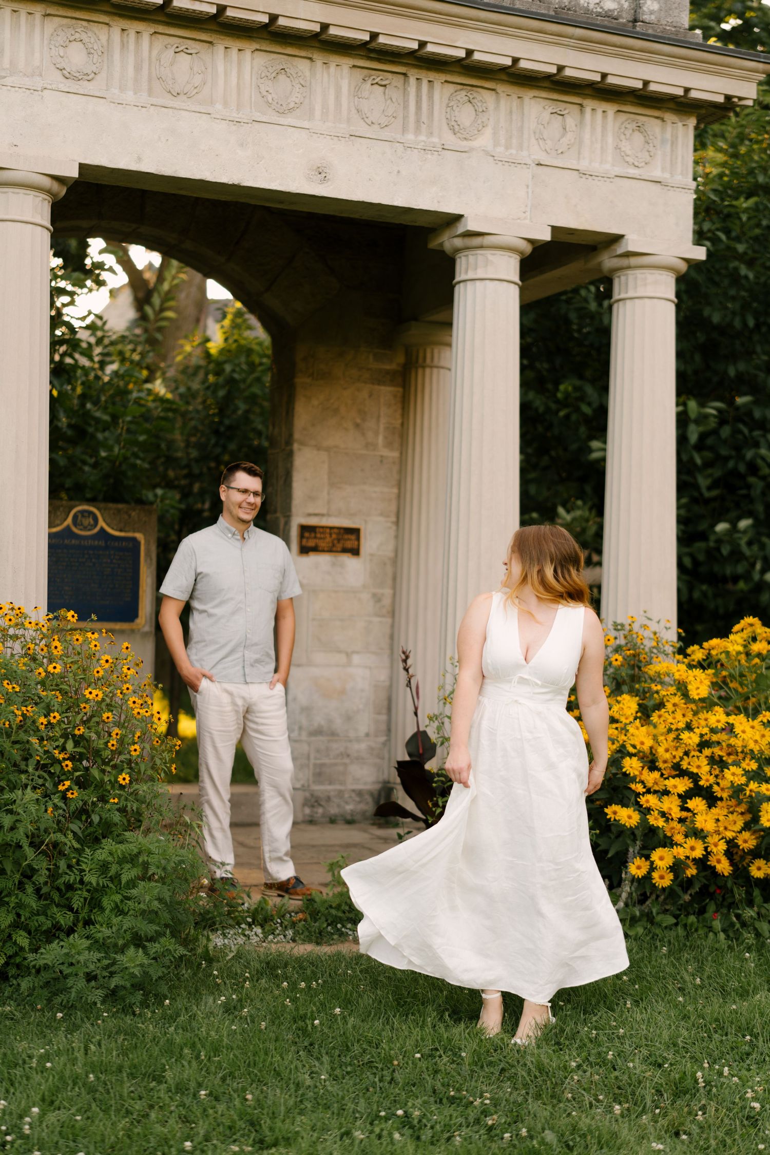 Woman in flowing white dress walking toward man standing by stone columned structure with yellow flowers.