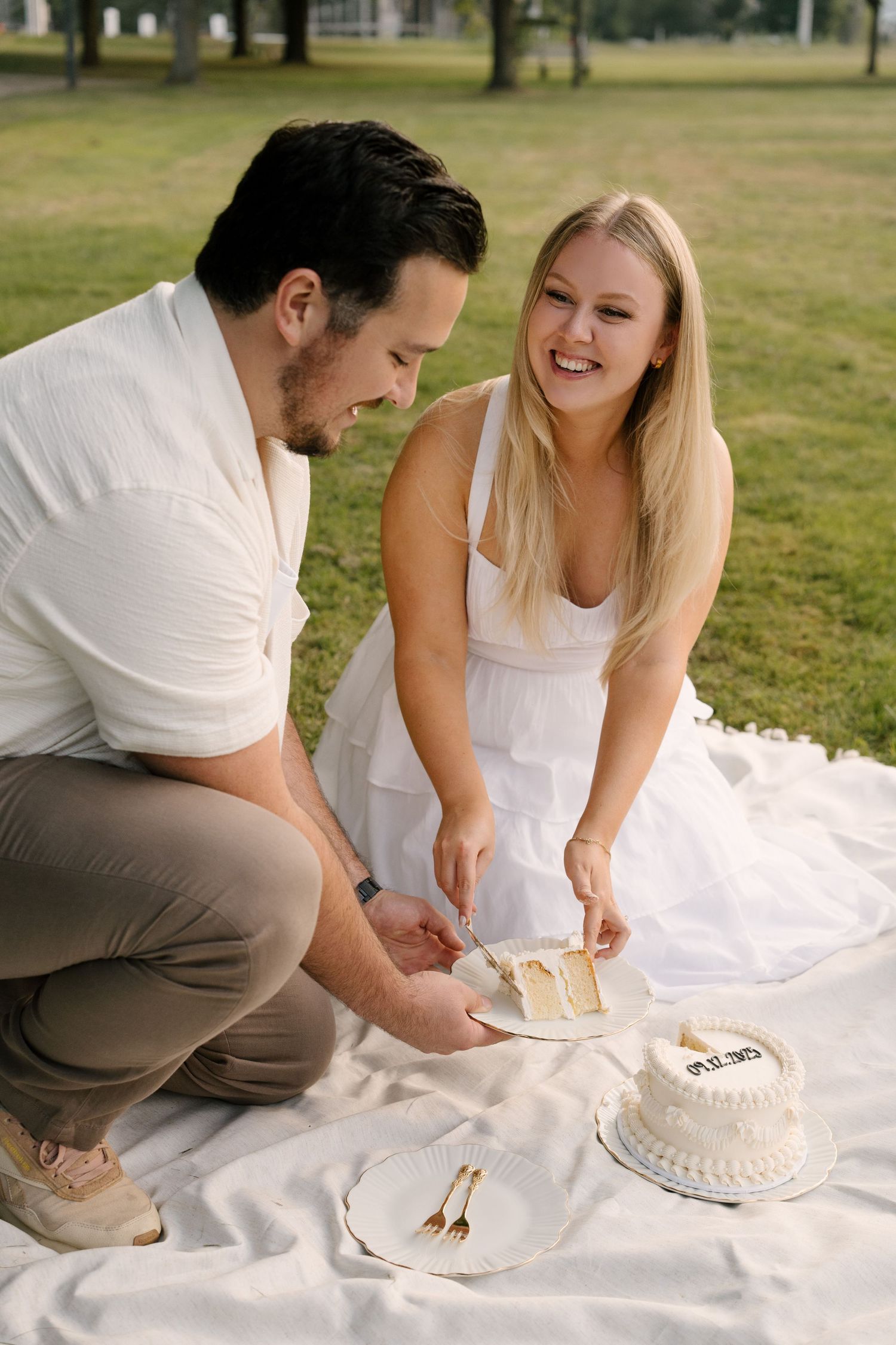 Happy couple cutting and serving slices of a white celebration cake on a picnic blanket in a park.