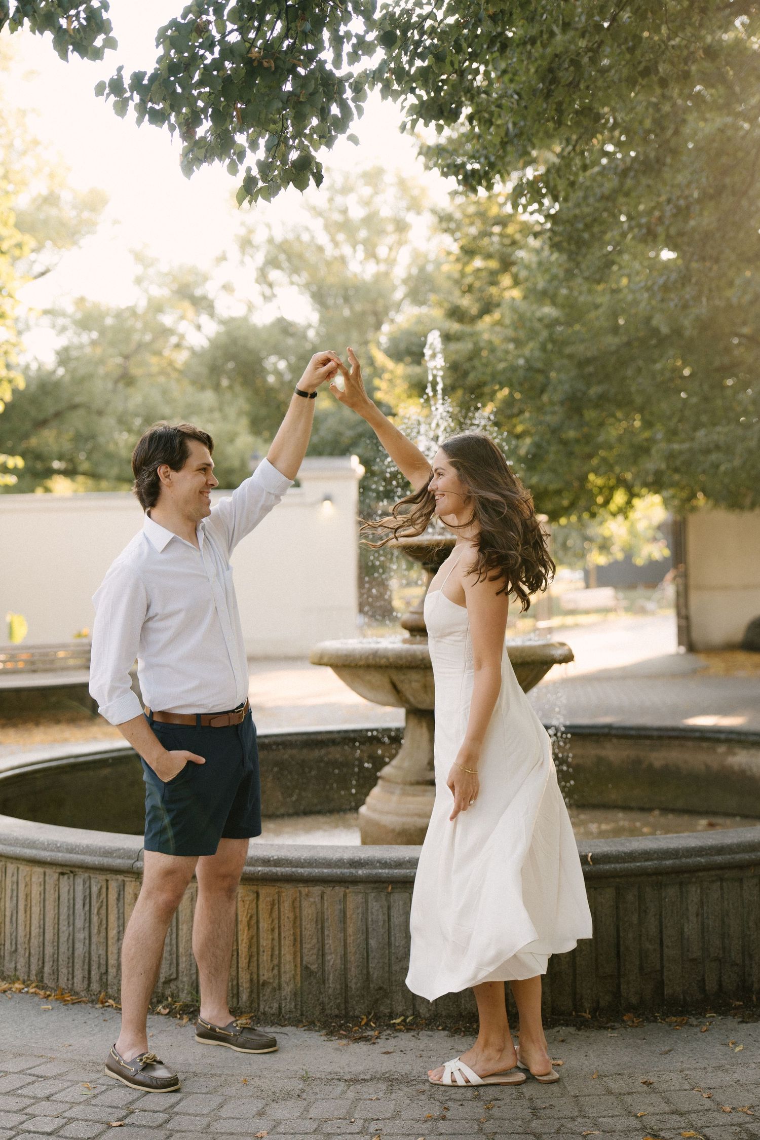 Couple dancing playfully near a stone fountain in a sunlit park with lush green trees.