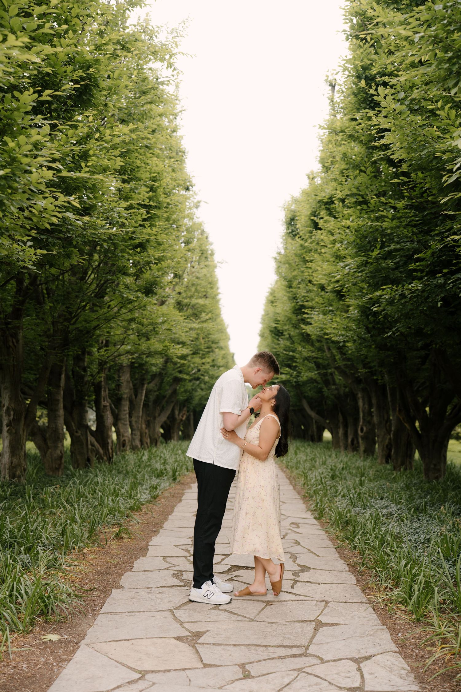 Couple sharing a tender kiss on a stone pathway lined with tall green trees.