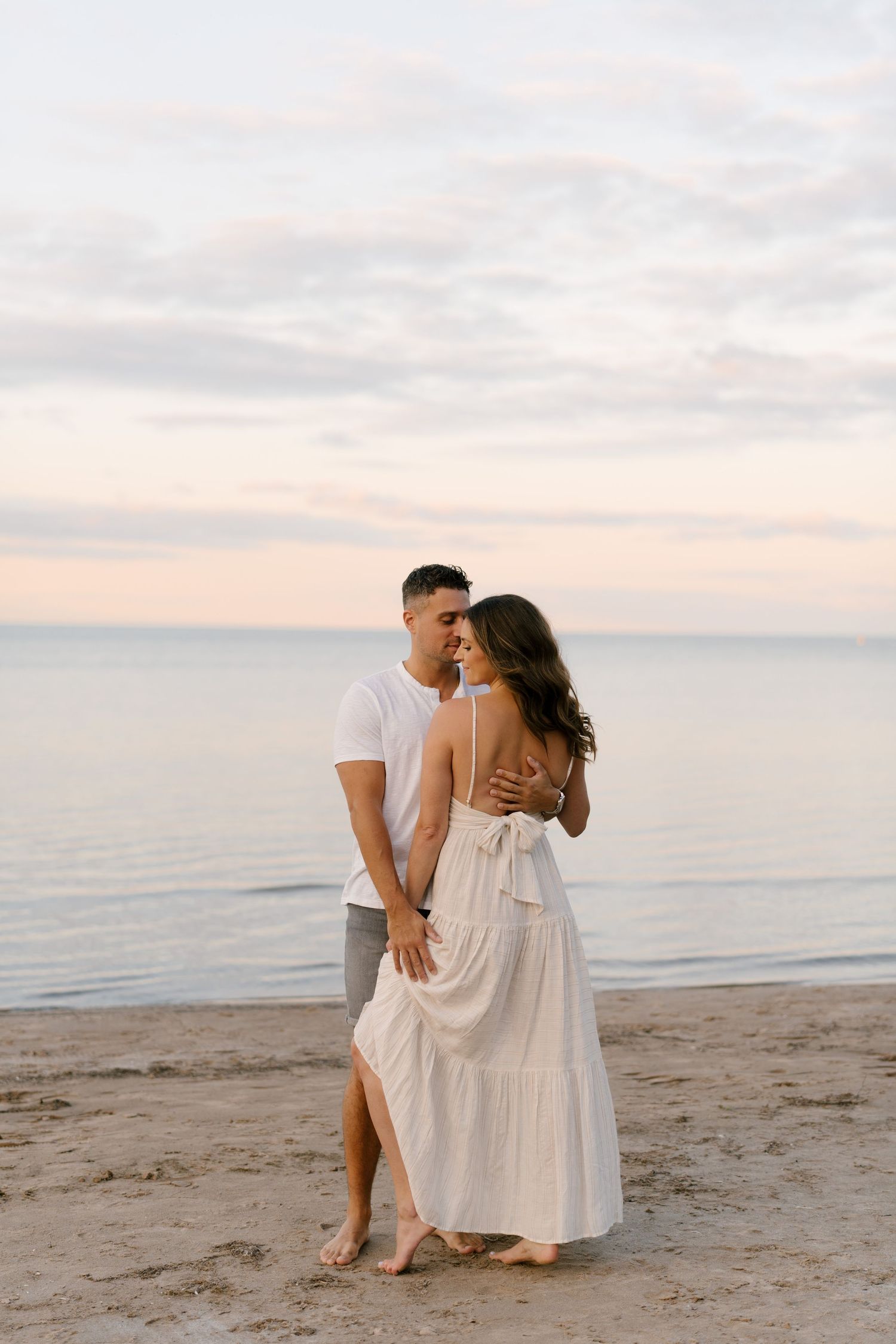 Couple embracing on a sandy beach at dusk, woman in white backless dress, man in white t-shirt.