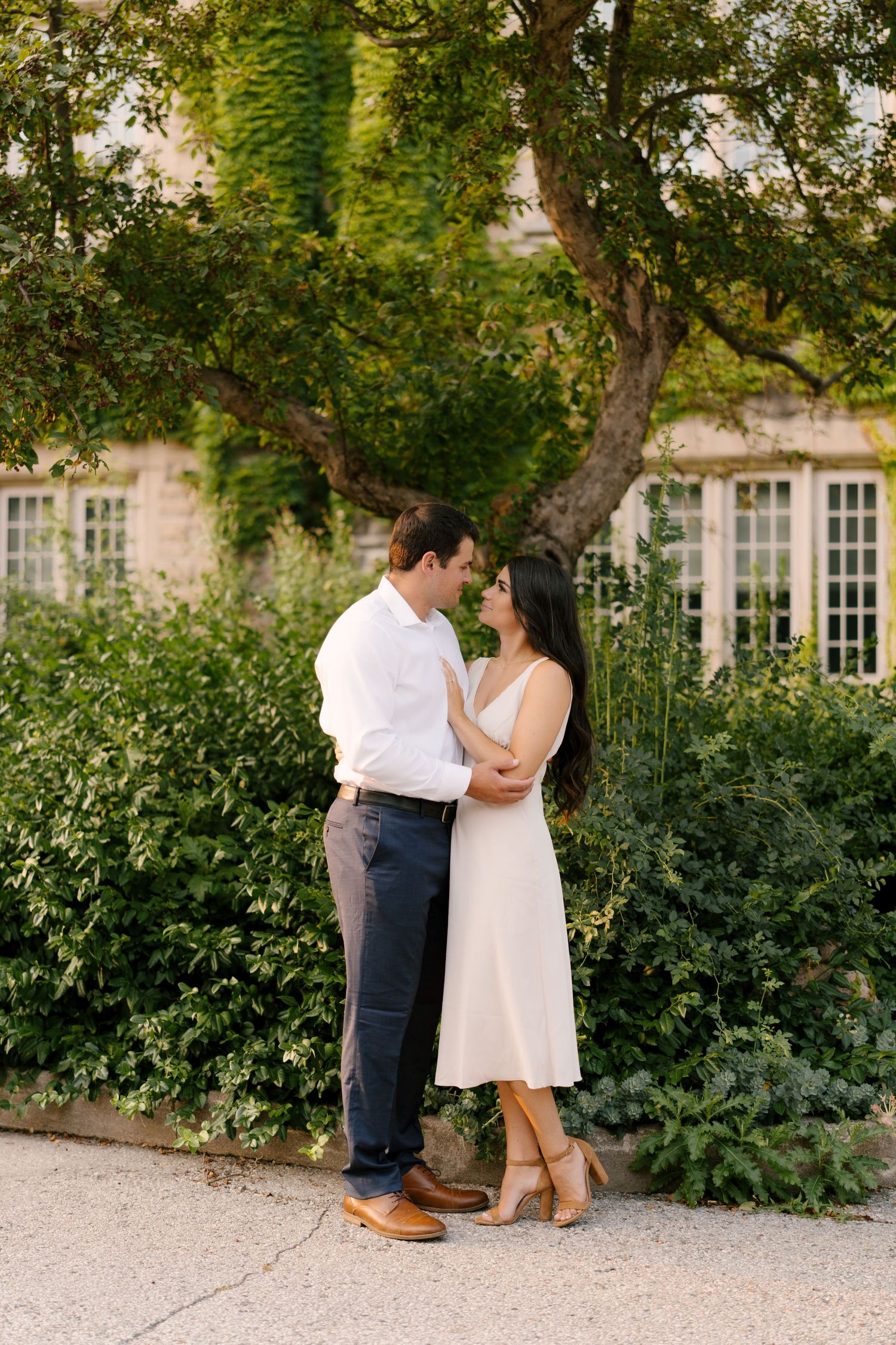 Couple gazing at each other tenderly in front of ivy-covered building surrounded by lush greenery.