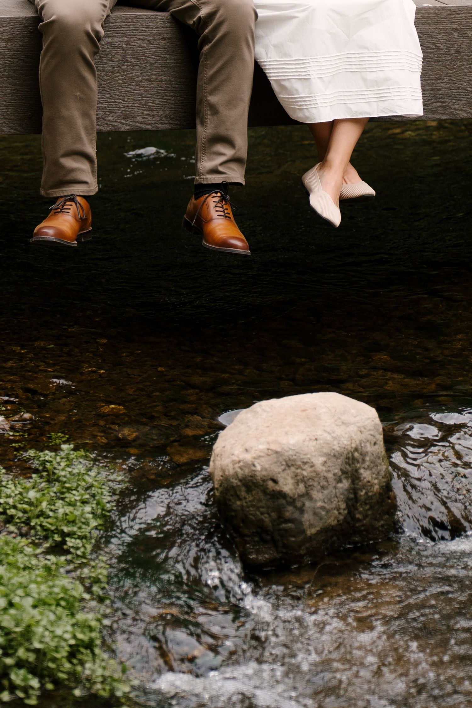 Close-up of couple's feet dangling over a rocky stream, one wearing brown leather dress shoes.