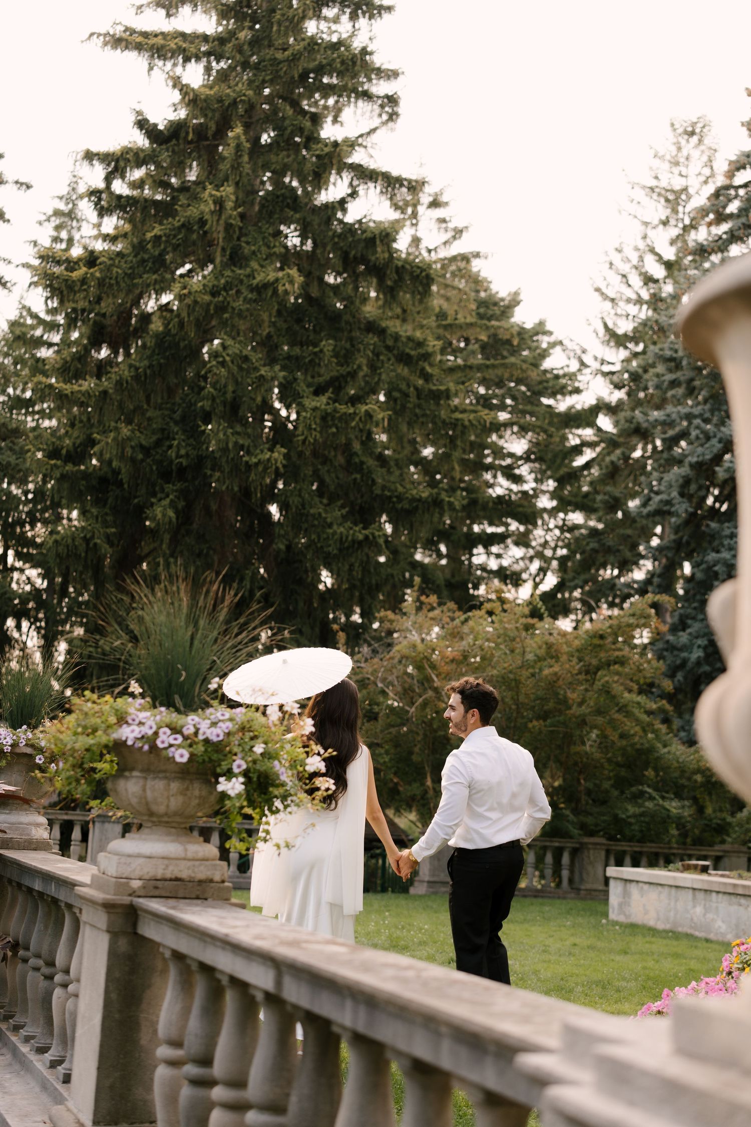 Elegant couple holding hands on a stone balustrade terrace surrounded by lush evergreen trees during a wedding photoshoot.
