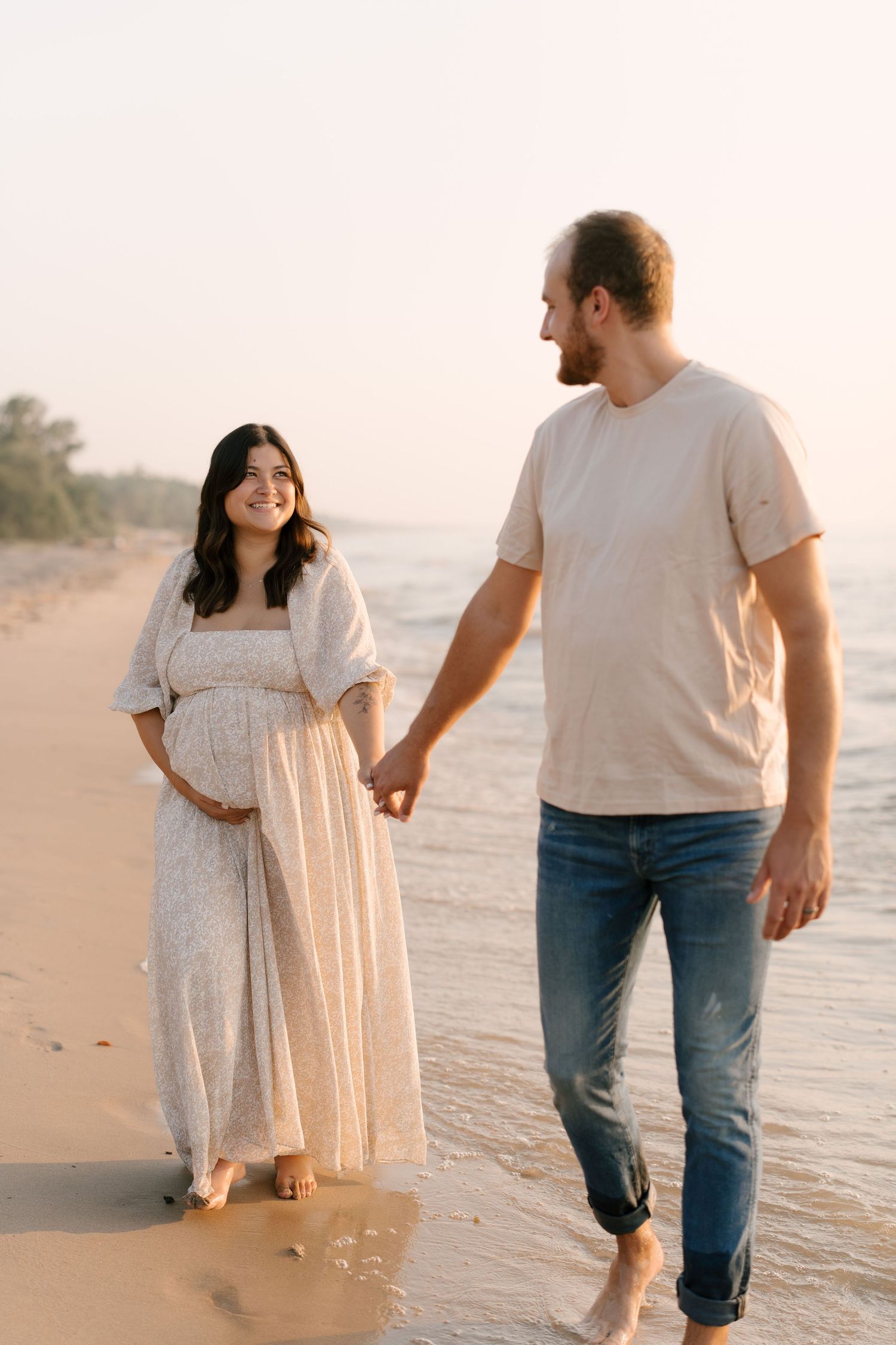 Smiling pregnant woman in white dress holds baby bump while walking with partner on beach.
