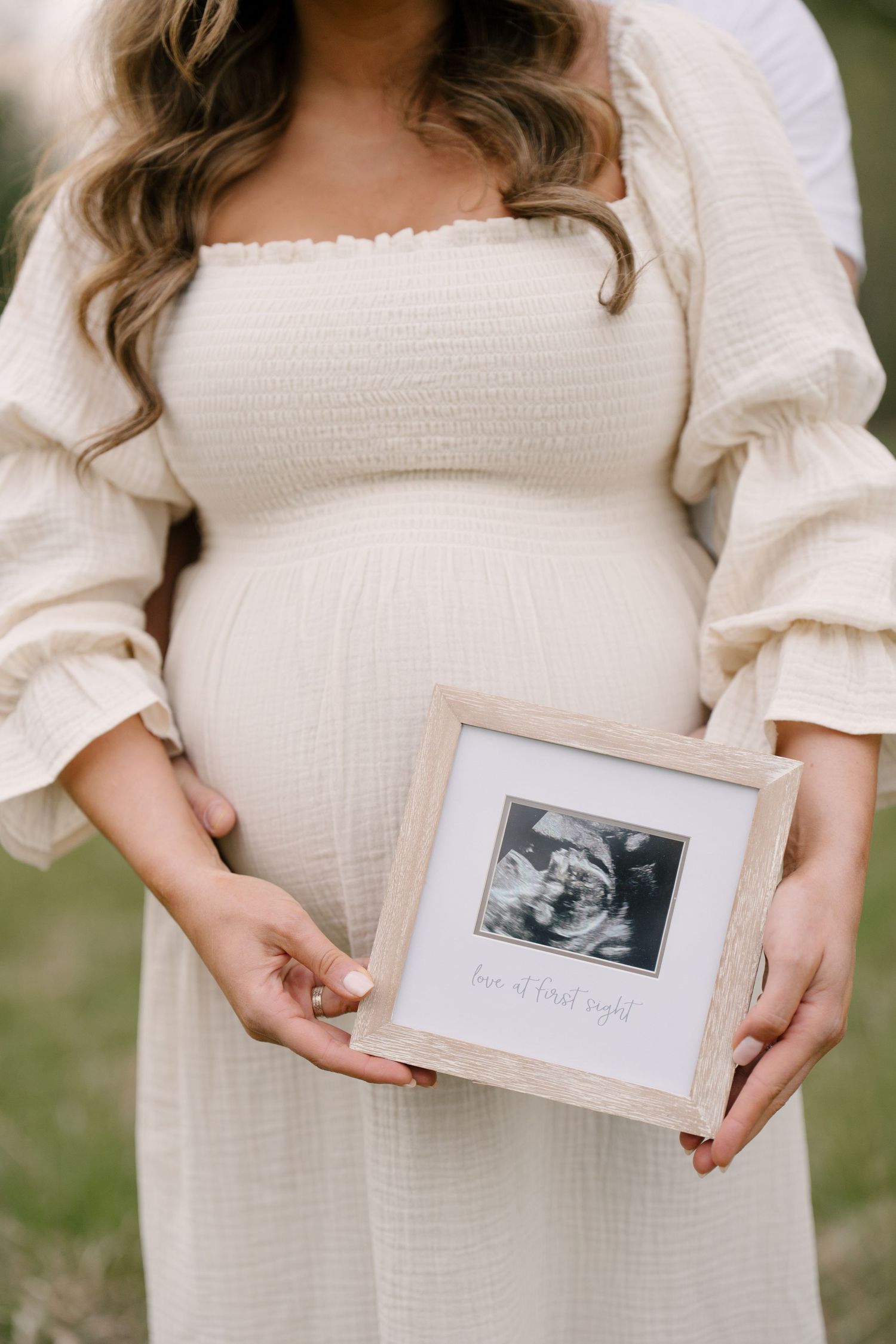 Pregnant woman in cream dress holding framed ultrasound photo outdoors.