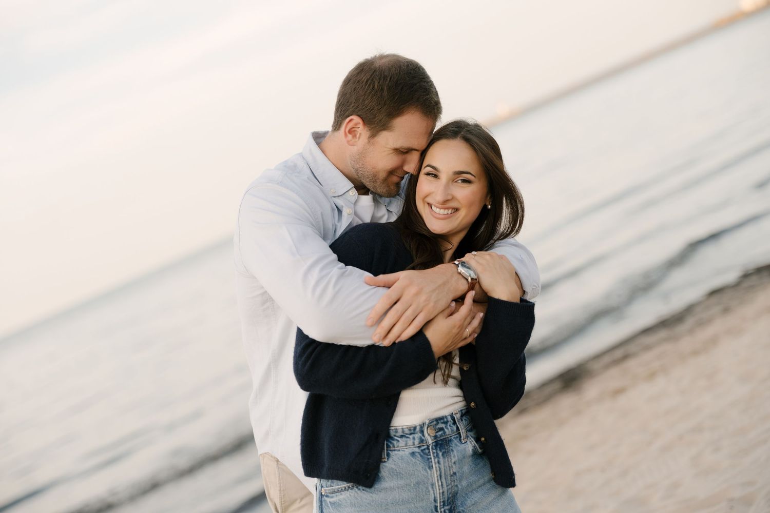 Couple sharing tender embrace on beach at sunset, woman in dark jacket and jeans smiling warmly.