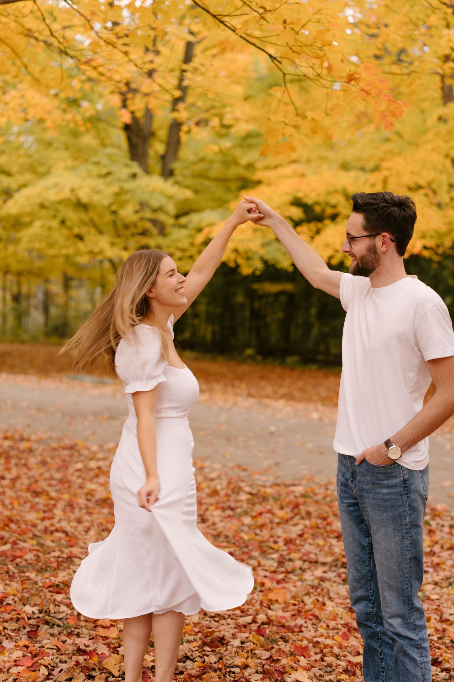 A couple dances together on a leaf-covered path surrounded by vibrant yellow autumn trees.