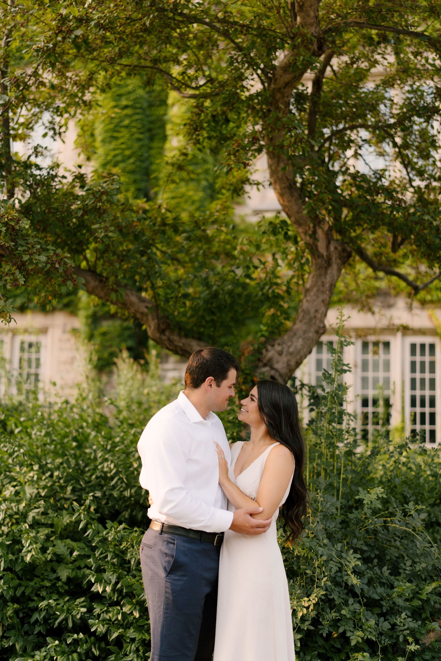 Couple embracing and smiling at each other before ivy-covered building in warm evening light.
