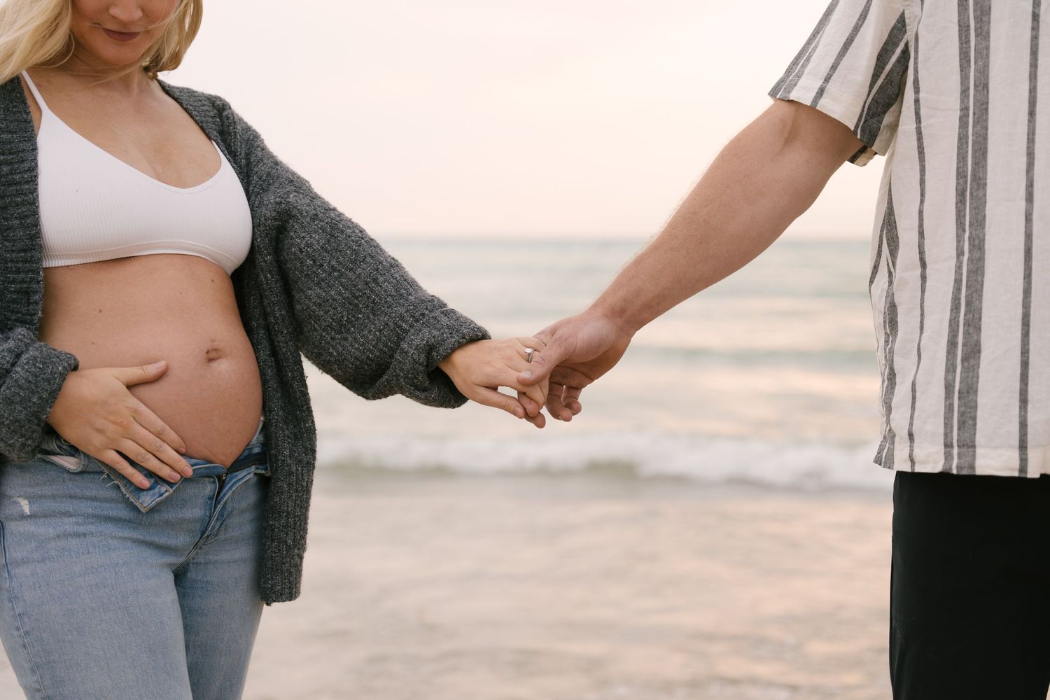 Close-up of pregnant couple holding hands on beach, woman cradling bare baby bump.