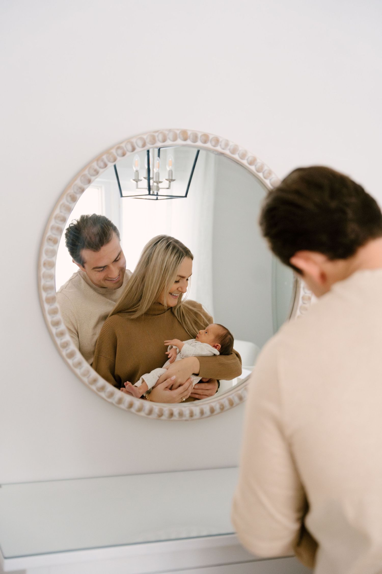 Couple with newborn baby reflected in round decorative mirror in bright white nursery room.