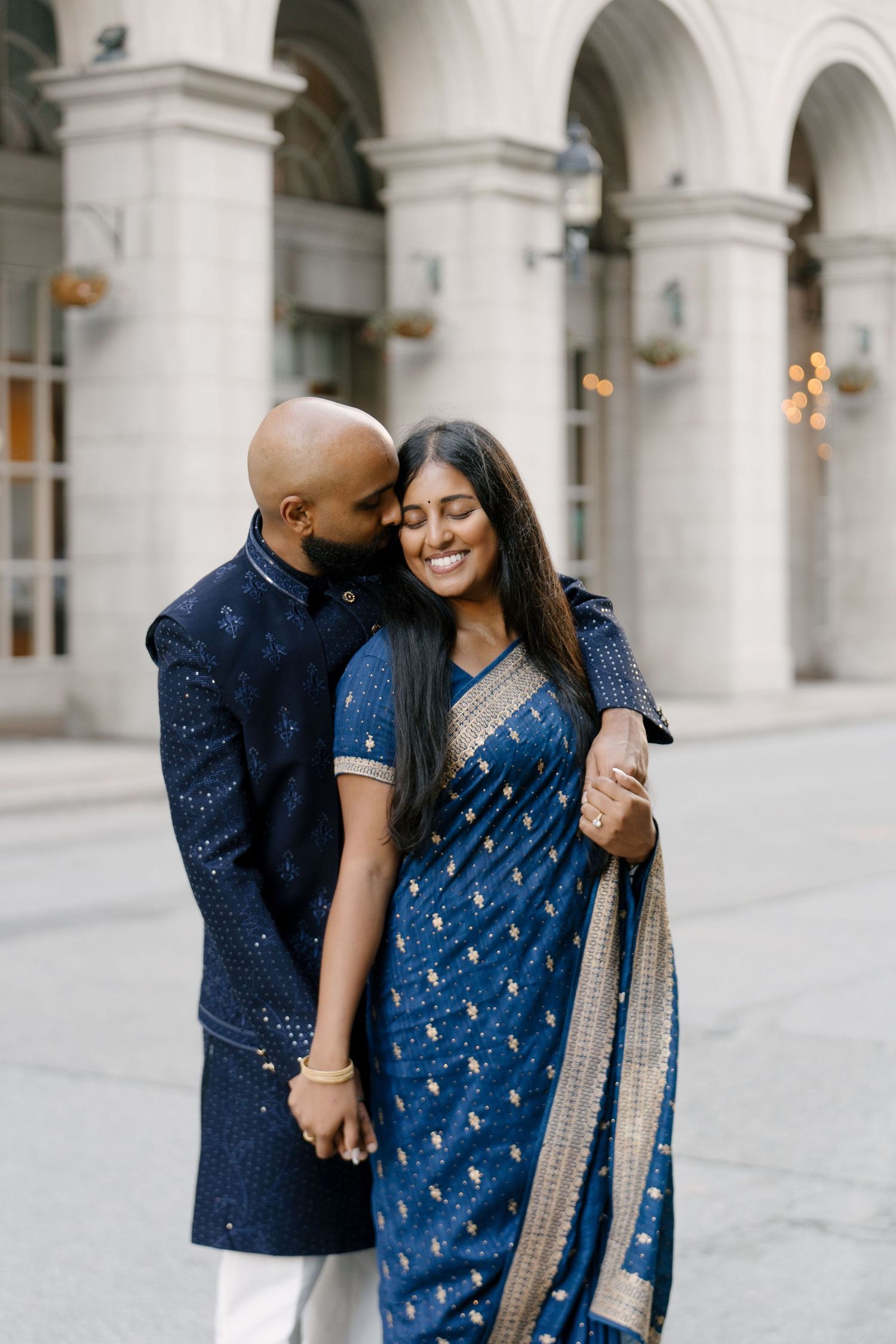Indian couple in blue traditional attire share a tender moment outside a classical building.