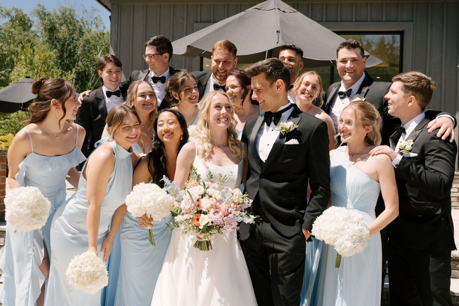 Bride and groom surrounded by laughing wedding party in light blue dresses and black tuxedos at Roseville Estates wedding venue.