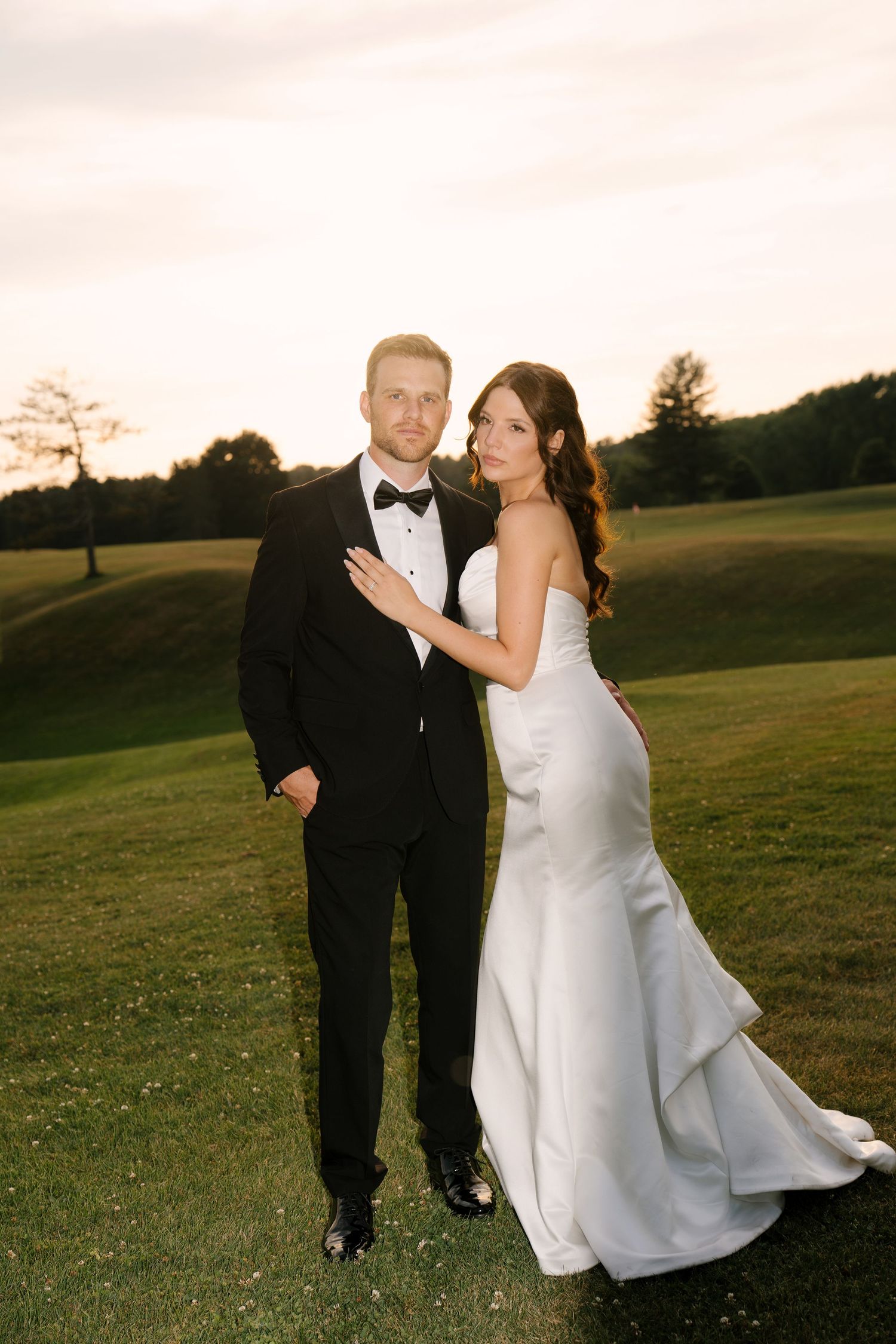 Bride in a fitted white gown embracing groom in a black tuxedo at golden hour outdoors.