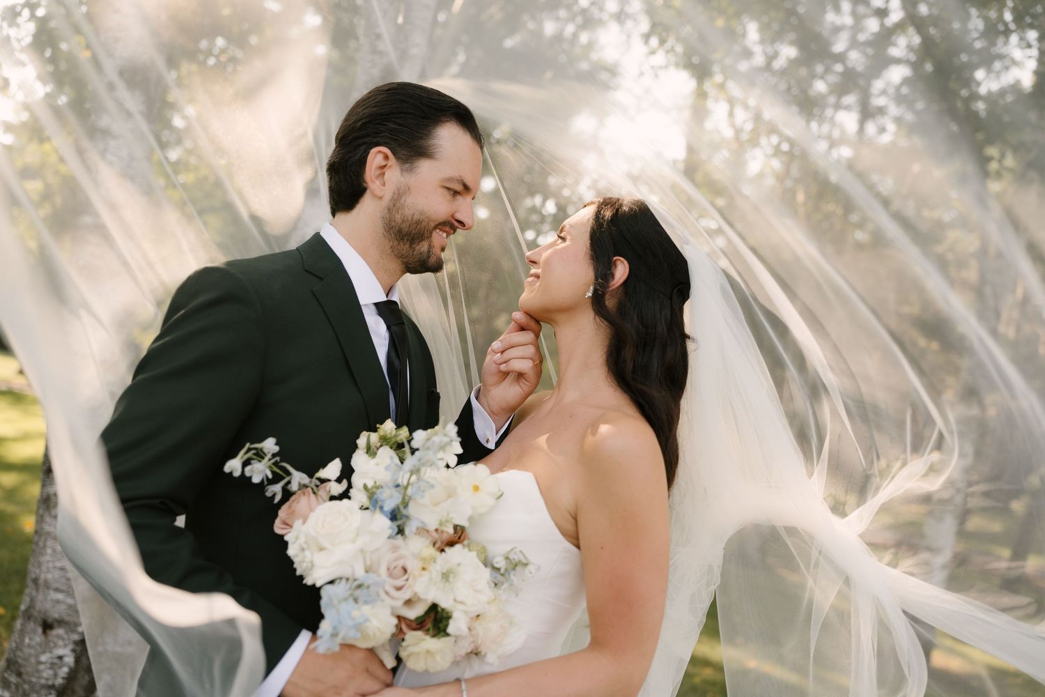 Bride and groom smiling under flowing veil outdoors, bride holding blue and white bouquet in romantic wedding portrait.