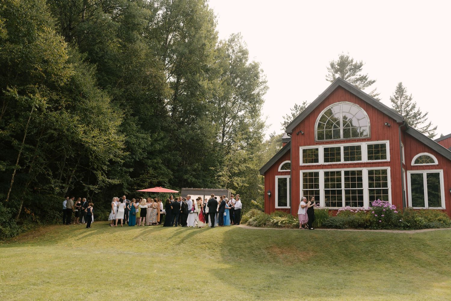 Wedding guests gather on a green lawn outside a picturesque red barn venue surrounded by tall trees.