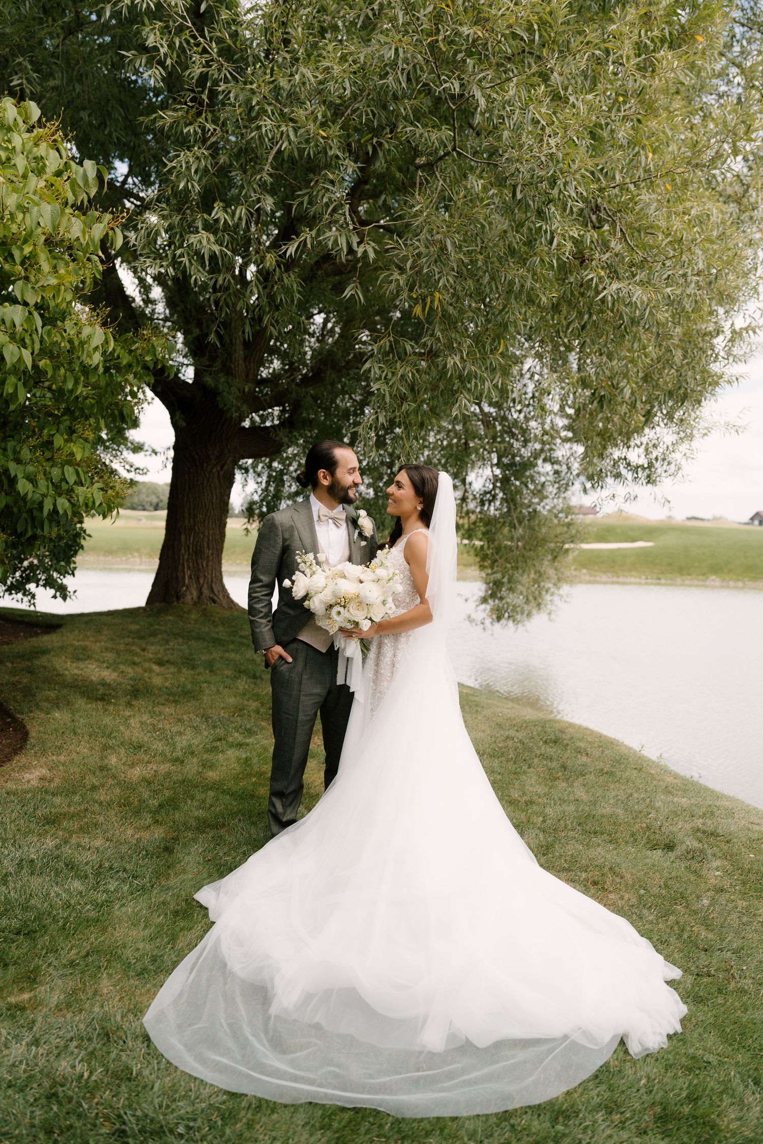 Bride and groom share a romantic moment under a willow tree by a lake, bride in a flowing white gown with veil.