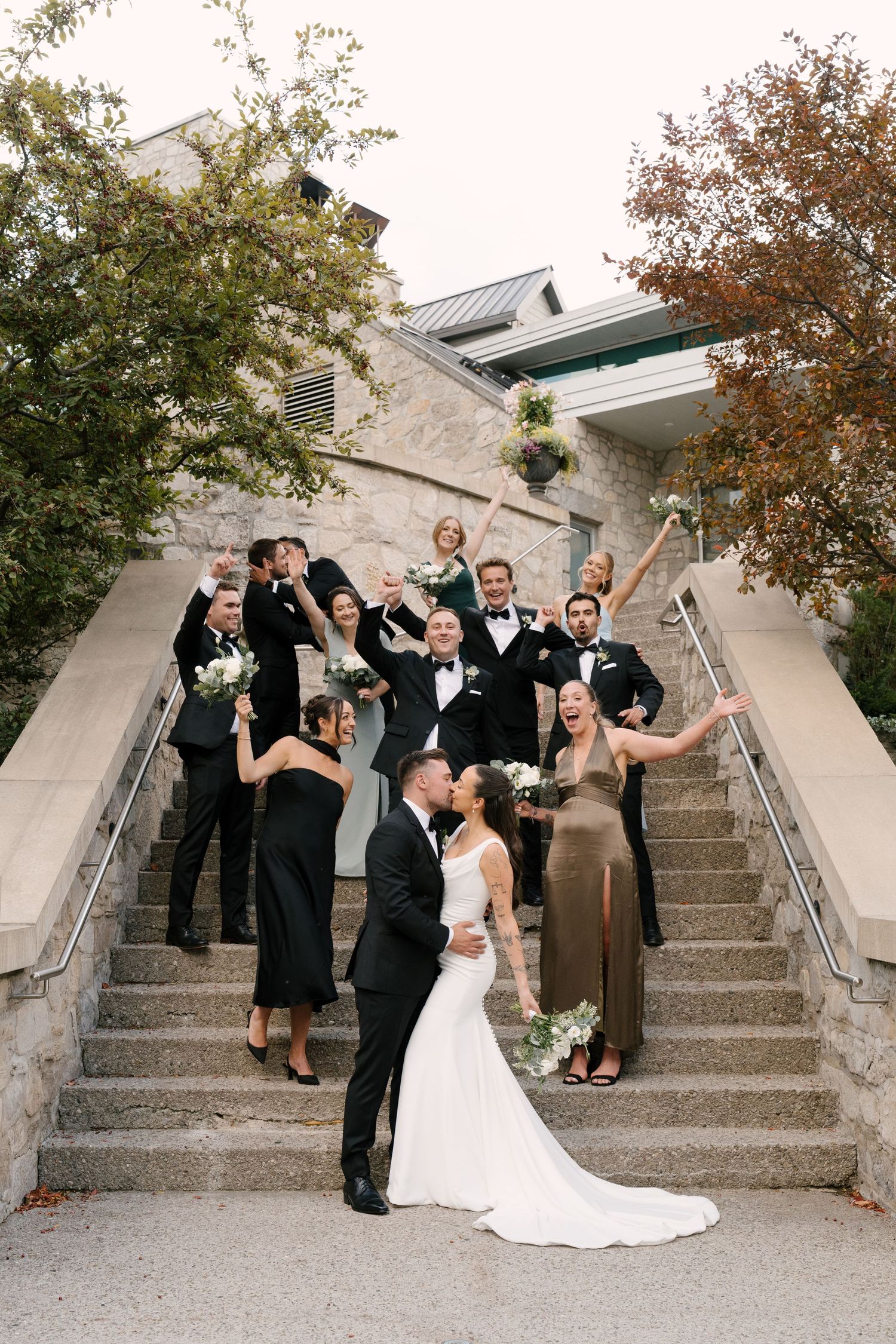 Wedding party celebrates on stone steps as bride and groom kiss in front of a historic Cambridge Mill venue.