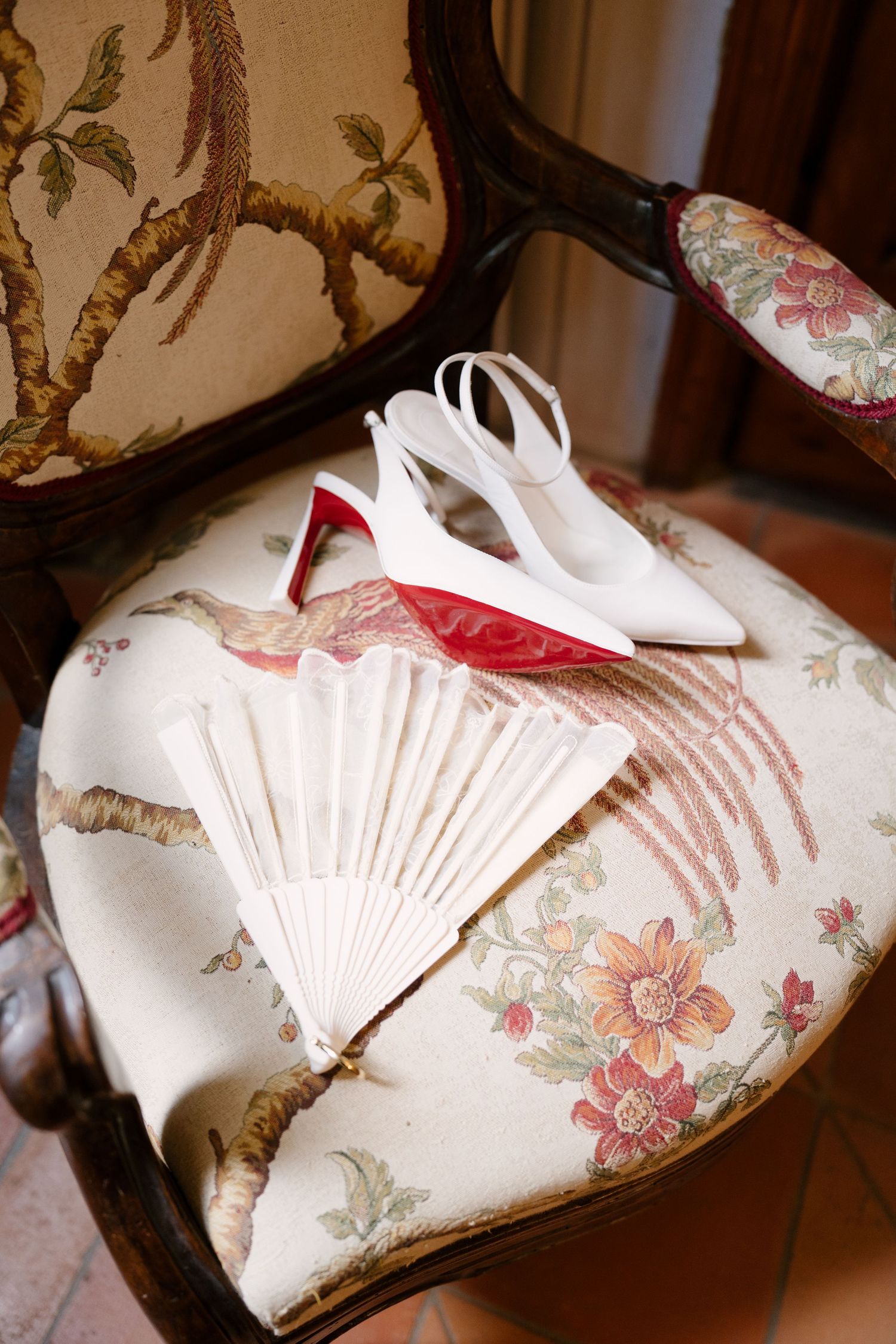 White slingback bridal heels with red soles and a folded white fan resting on a floral antique chair.