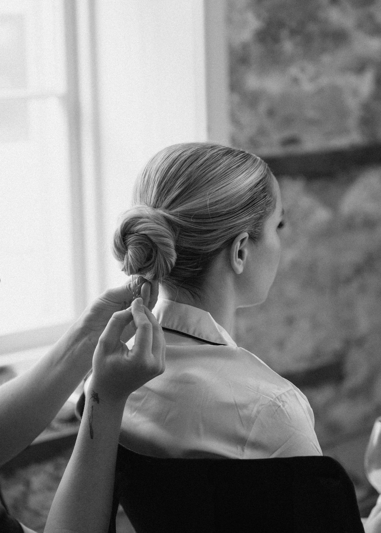 Black and white photo of bride's elegant low bun hairstyle being styled by a hairdresser near window.