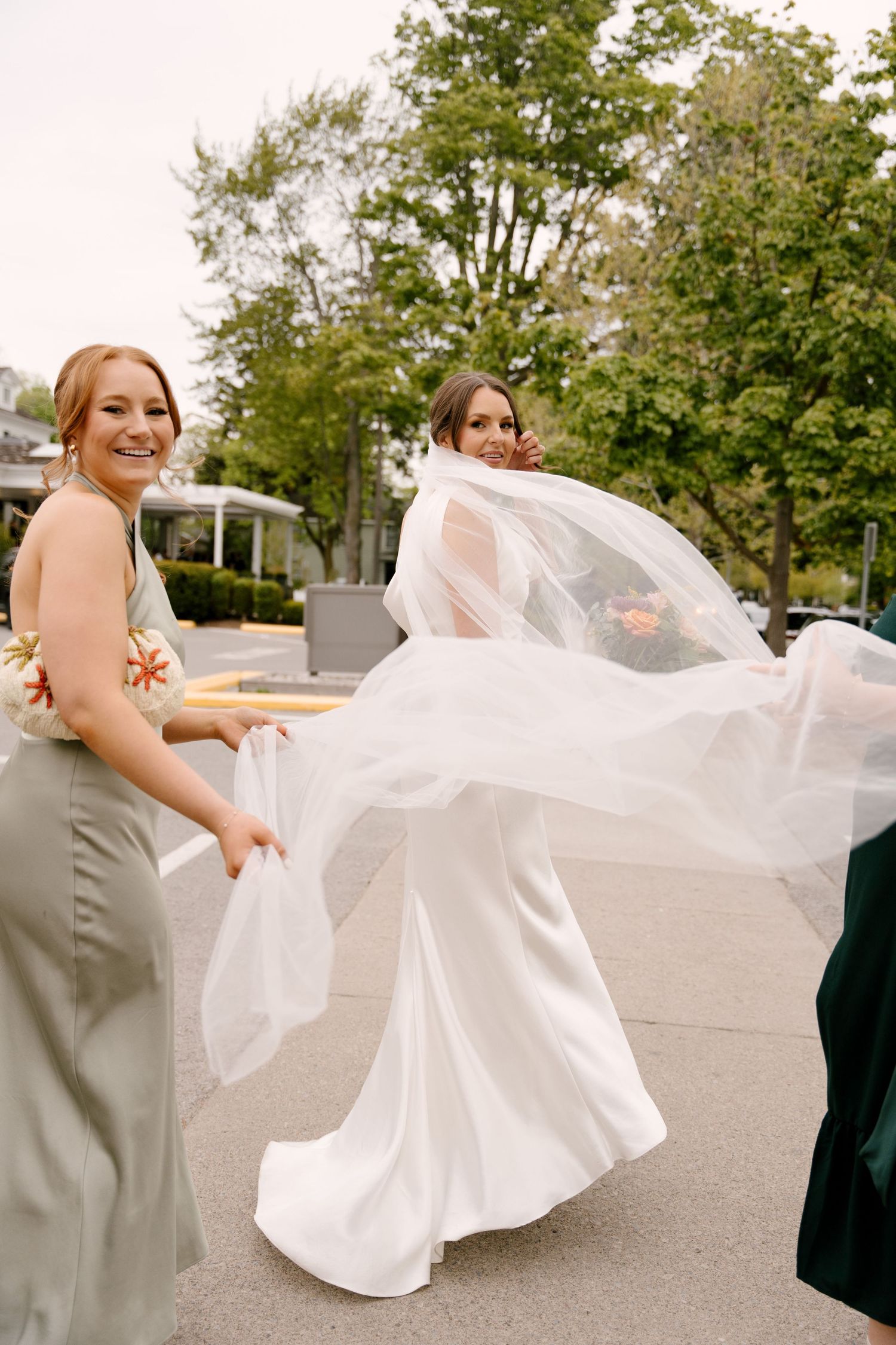 Bride twirls in white gown with veil flowing as bridesmaids assist outdoors on wedding day.