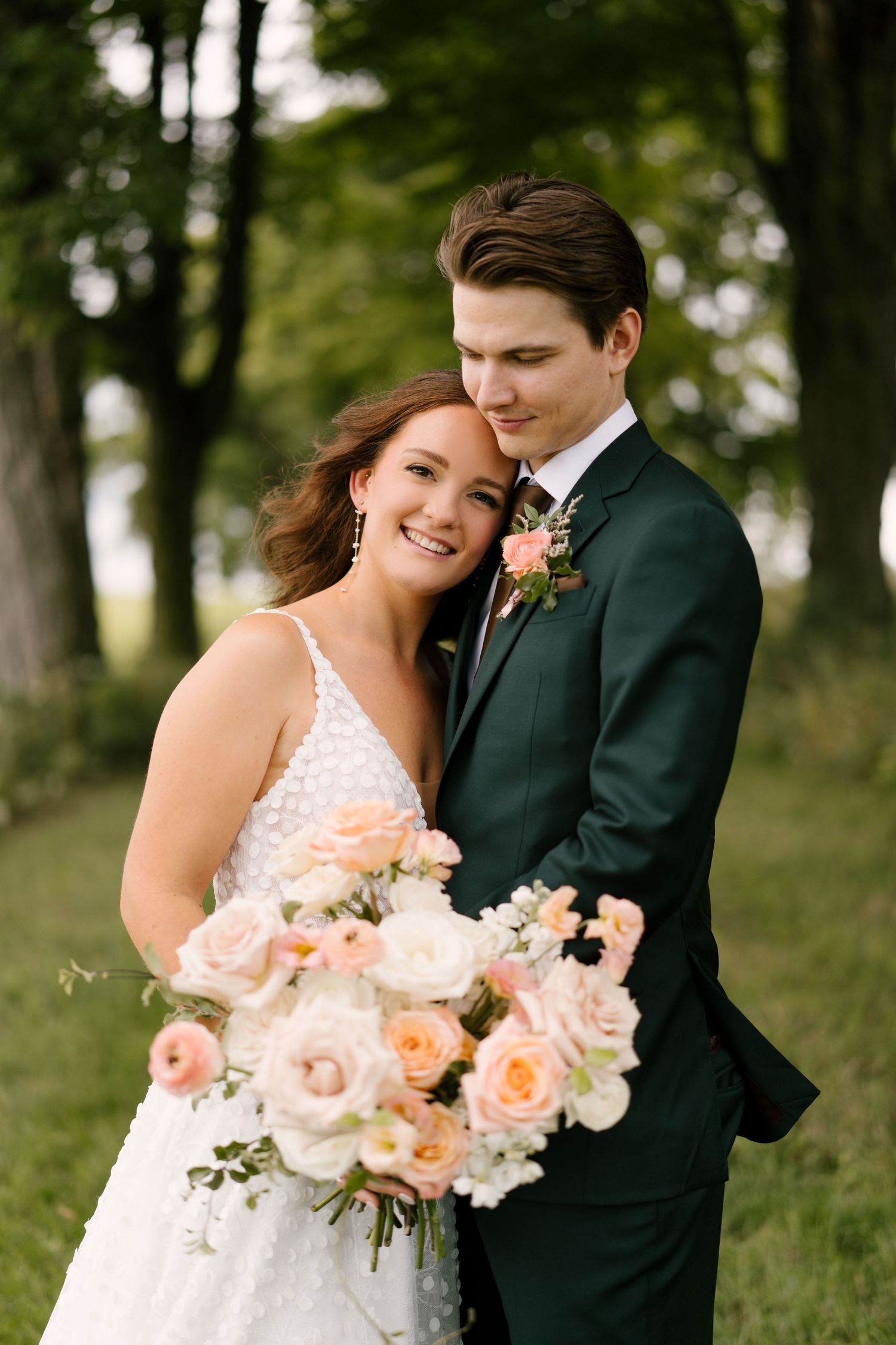 Groom tenderly embracing smiling bride holding peach and white rose bouquet among green trees.