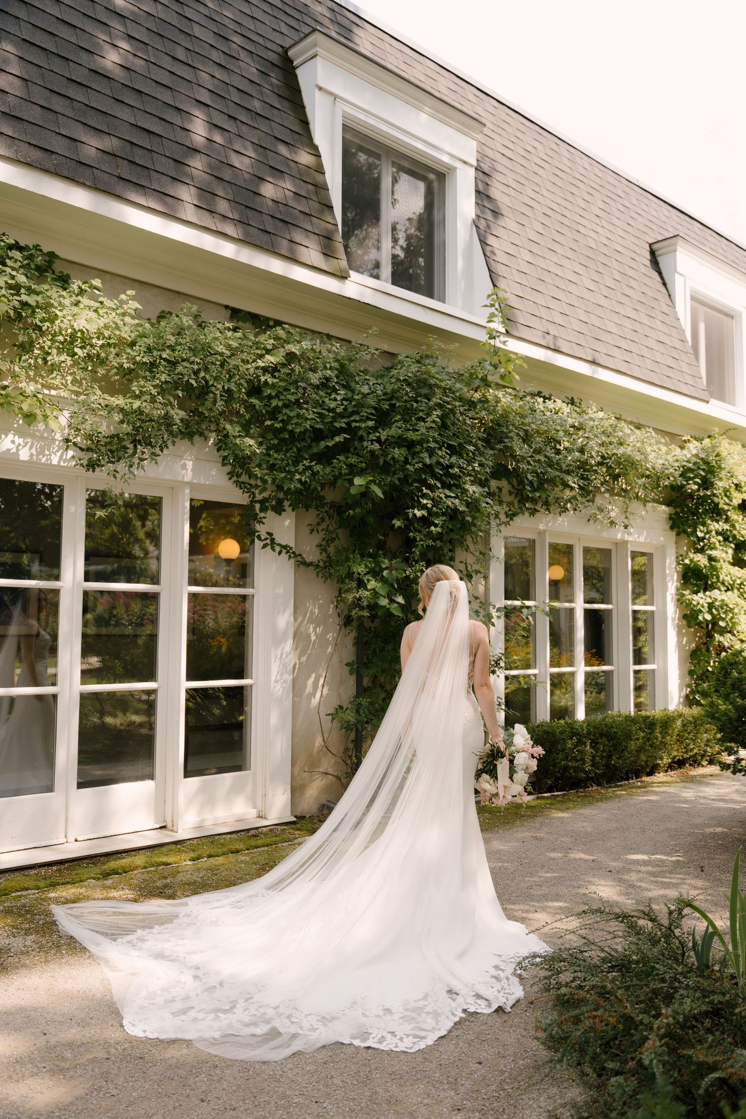 Bride in elegant lace wedding gown and cathedral veil stands before ivy-covered white Langdon Hall venue.
