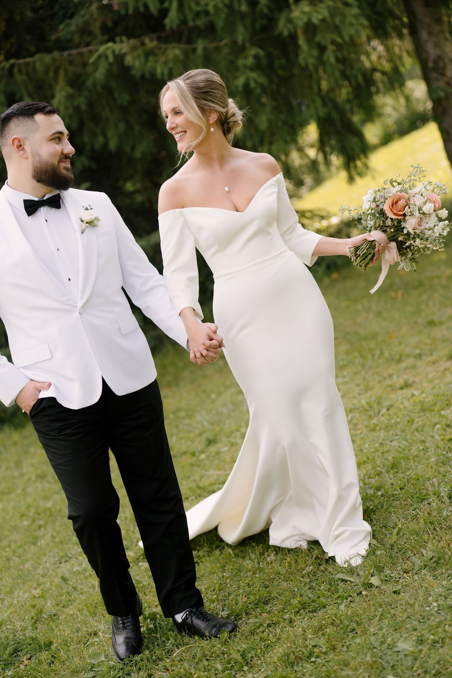 Bride and groom holding hands walking on green grass, bride in off-shoulder white gown holding bouquet, groom in white tuxedo.