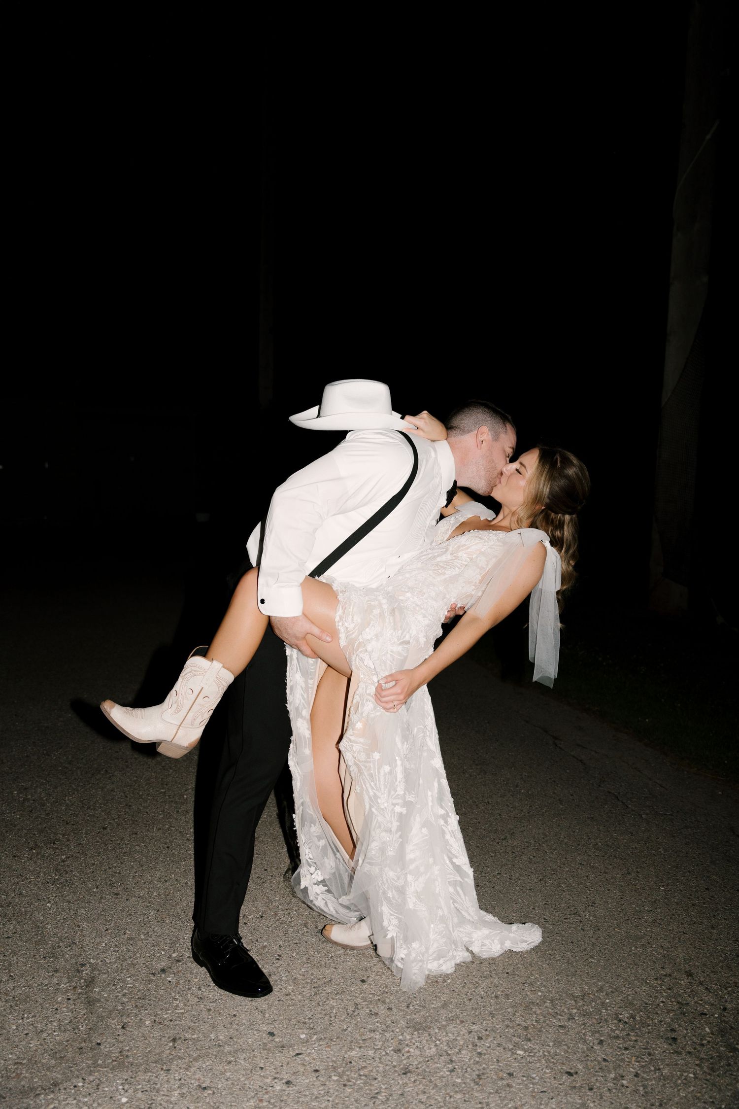Groom in black suit and white cowboy hat dips bride in lace wedding dress and white boots for a romantic kiss outside St Marys Venue.