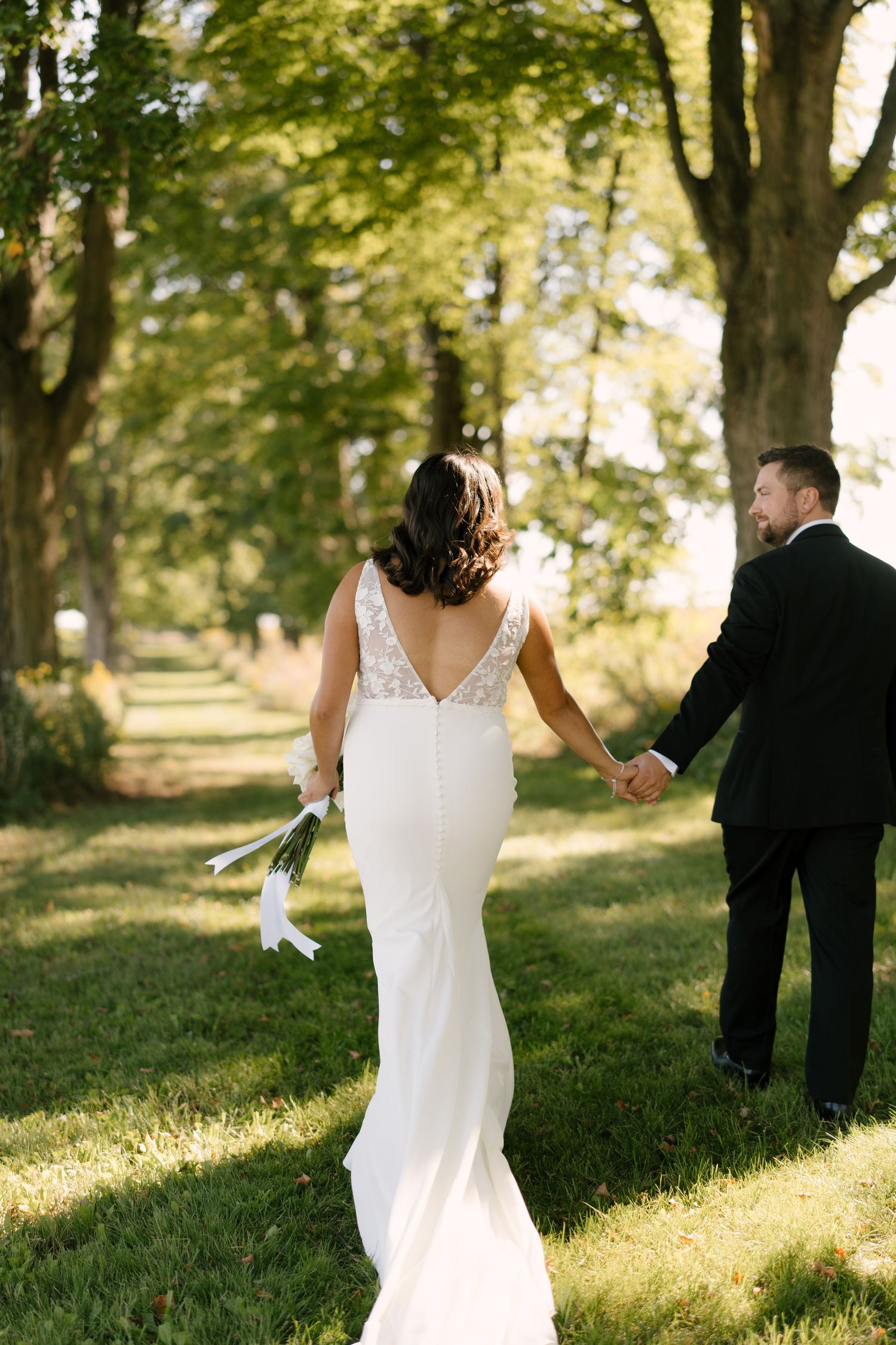Bride and groom walk hand in hand through tree-lined grassy path in outdoor wedding portrait.