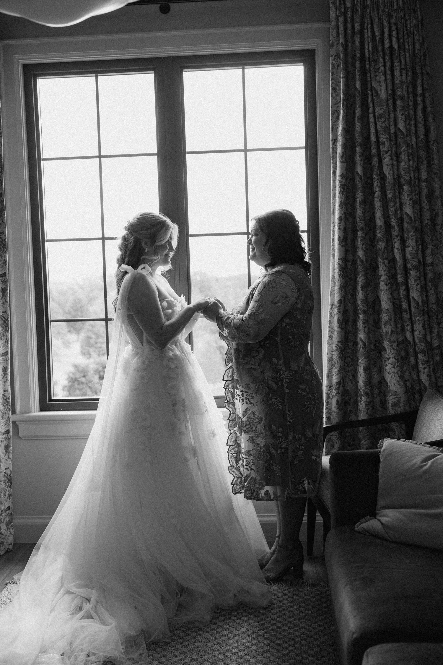 Black and white photo of bride and mother sharing tender moment by window before ceremony.
