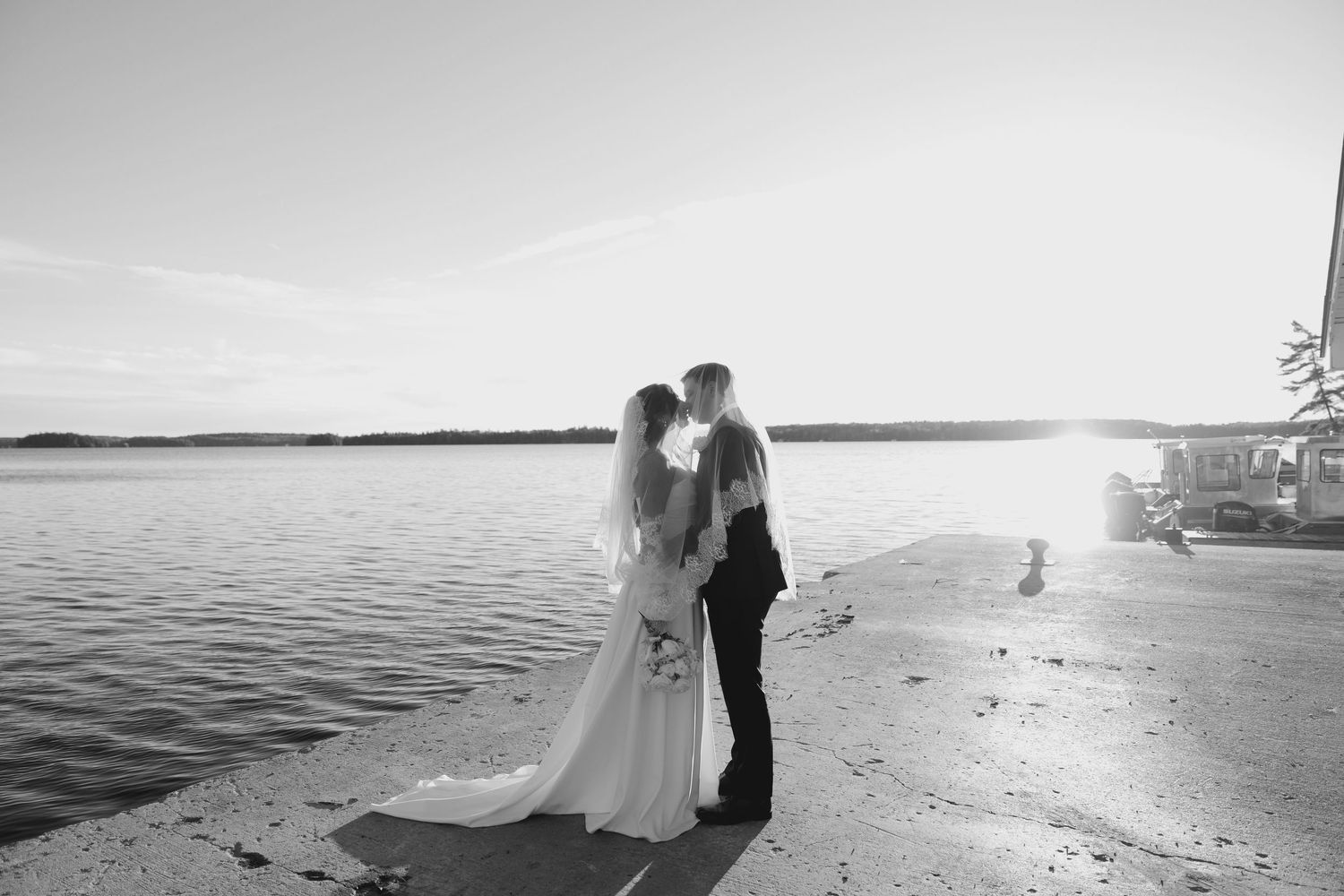 Black and white romantic photo of bride and groom kissing by the water at sunset.