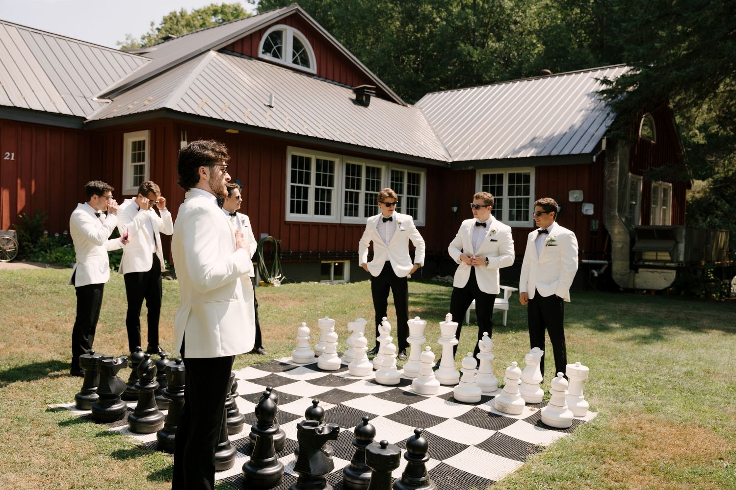 Groomsmen in white tuxedos playing giant outdoor chess on a black and white board near a red barn.