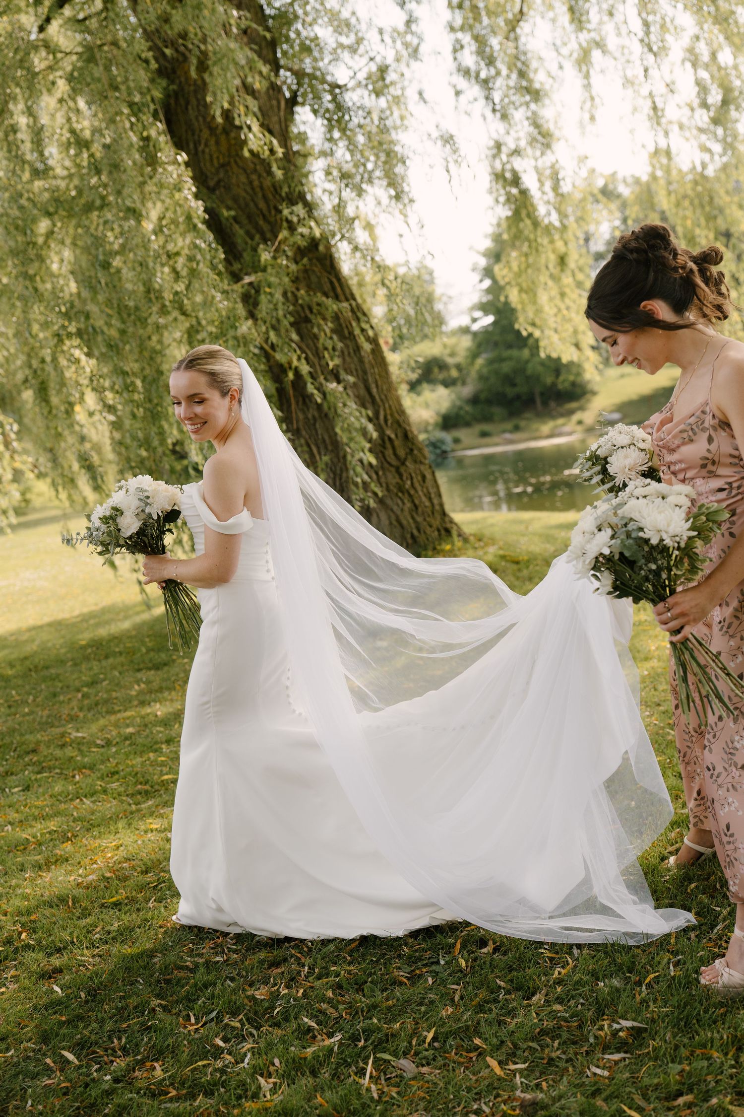 Bridesmaid helps arrange bride's cathedral veil and train in sunlit garden near willow tree.