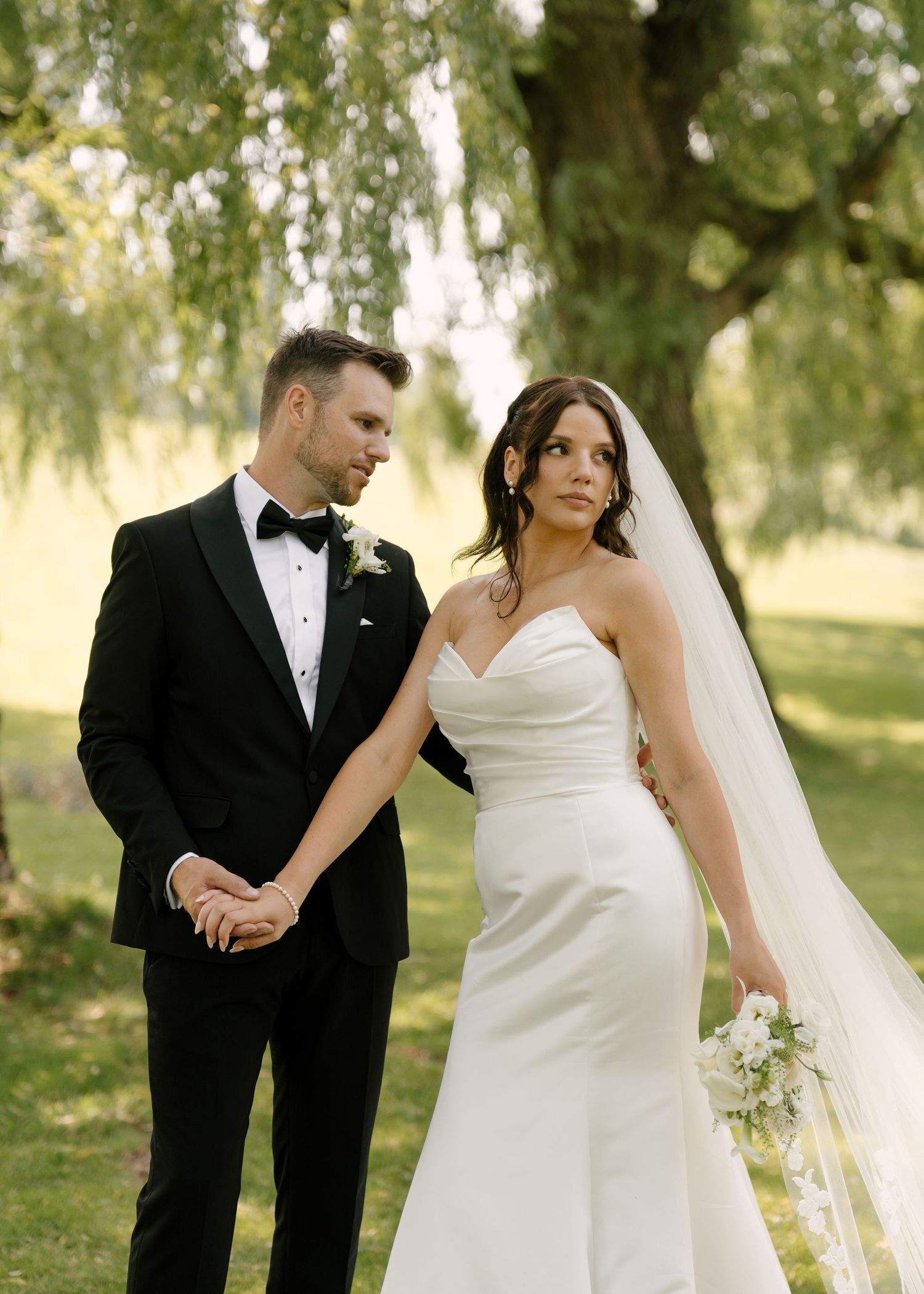 Bride in strapless white gown and groom in black tuxedo hold hands under a willow tree outdoors.