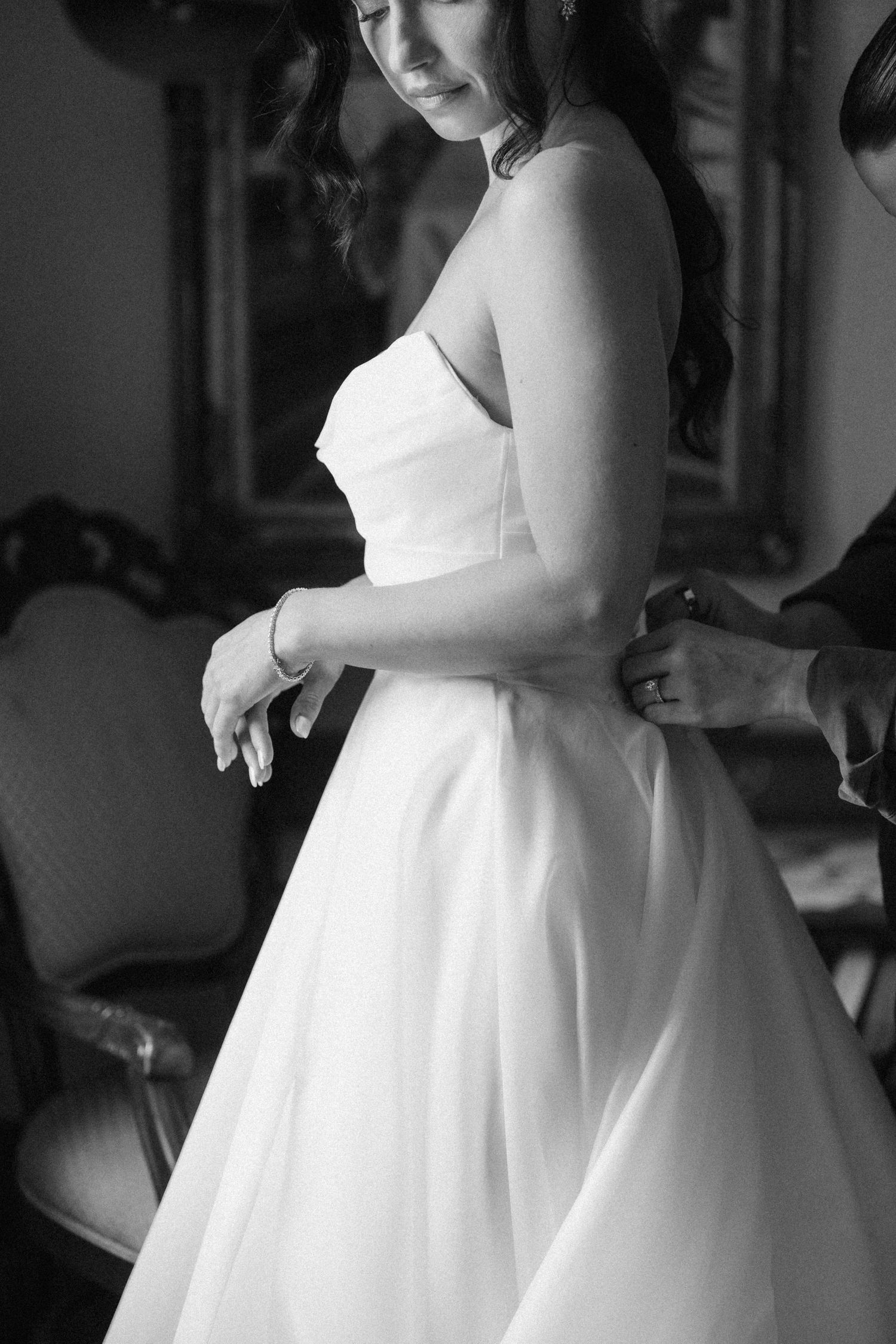 Black and white close-up of bride in strapless wedding dress being buttoned up before ceremony.