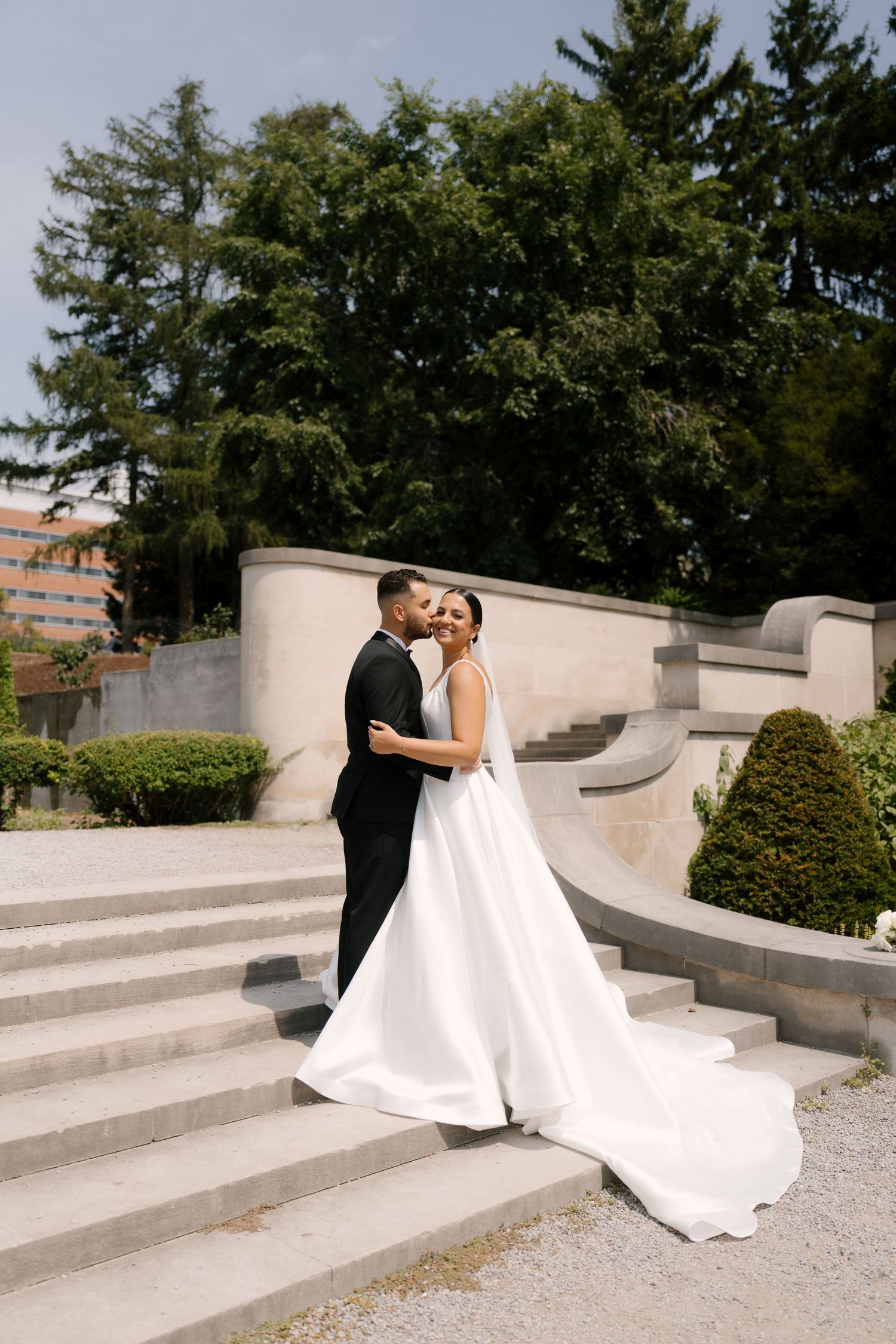 Bride and groom embracing on stone steps surrounded by manicured gardens and tall evergreen trees on wedding day at Parkwood Estates.