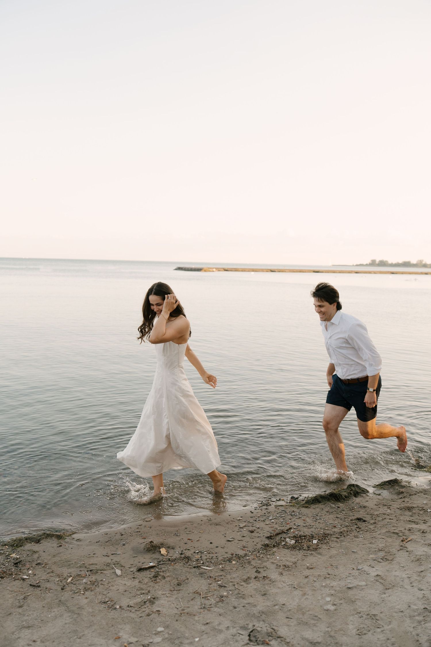 Couple running barefoot along a serene beach shoreline at sunset in casual attire.