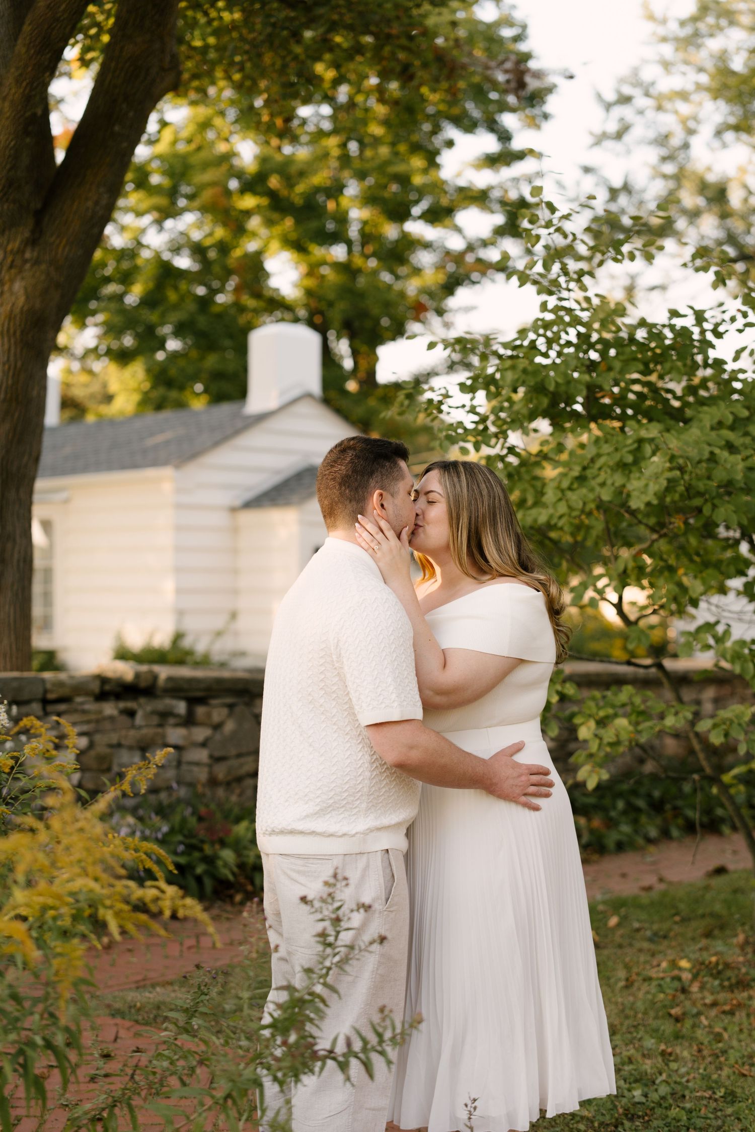 Couple sharing a romantic kiss in a sunlit garden with a white cottage and stone wall behind them.