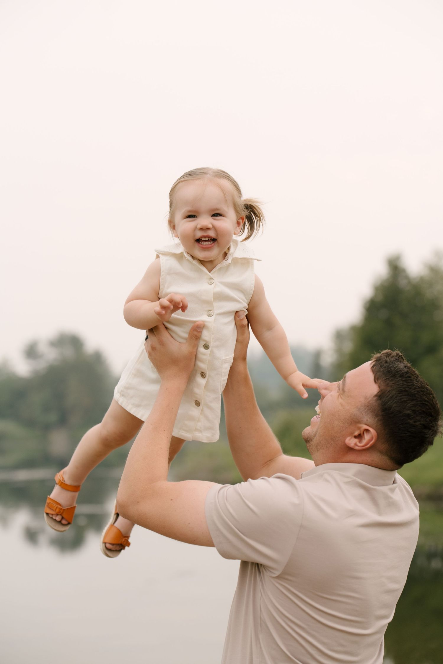Joyful father lifting smiling baby girl in the air near a peaceful lake.