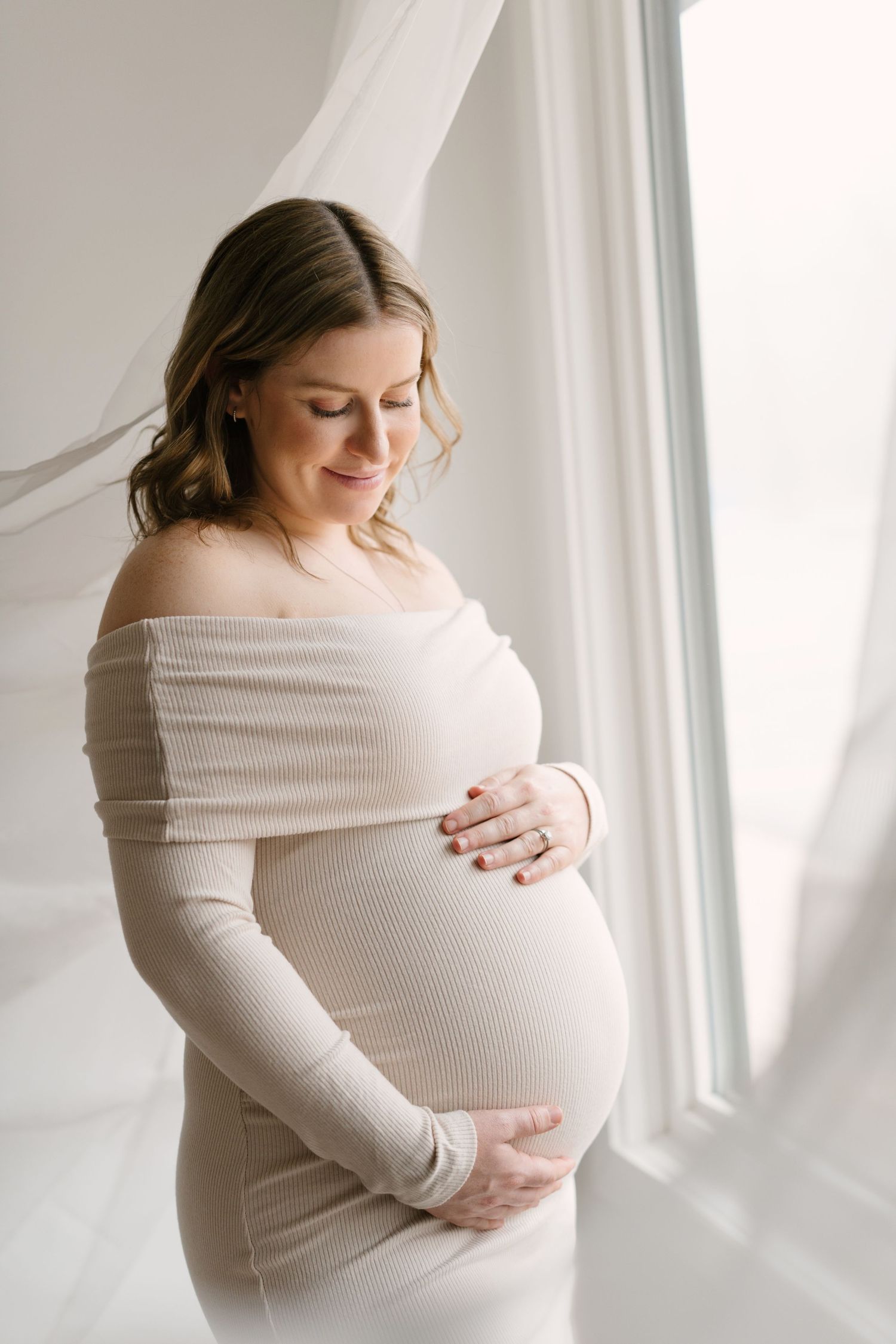 Smiling pregnant woman in cream off-shoulder ribbed dress cradling her baby bump near a bright window.